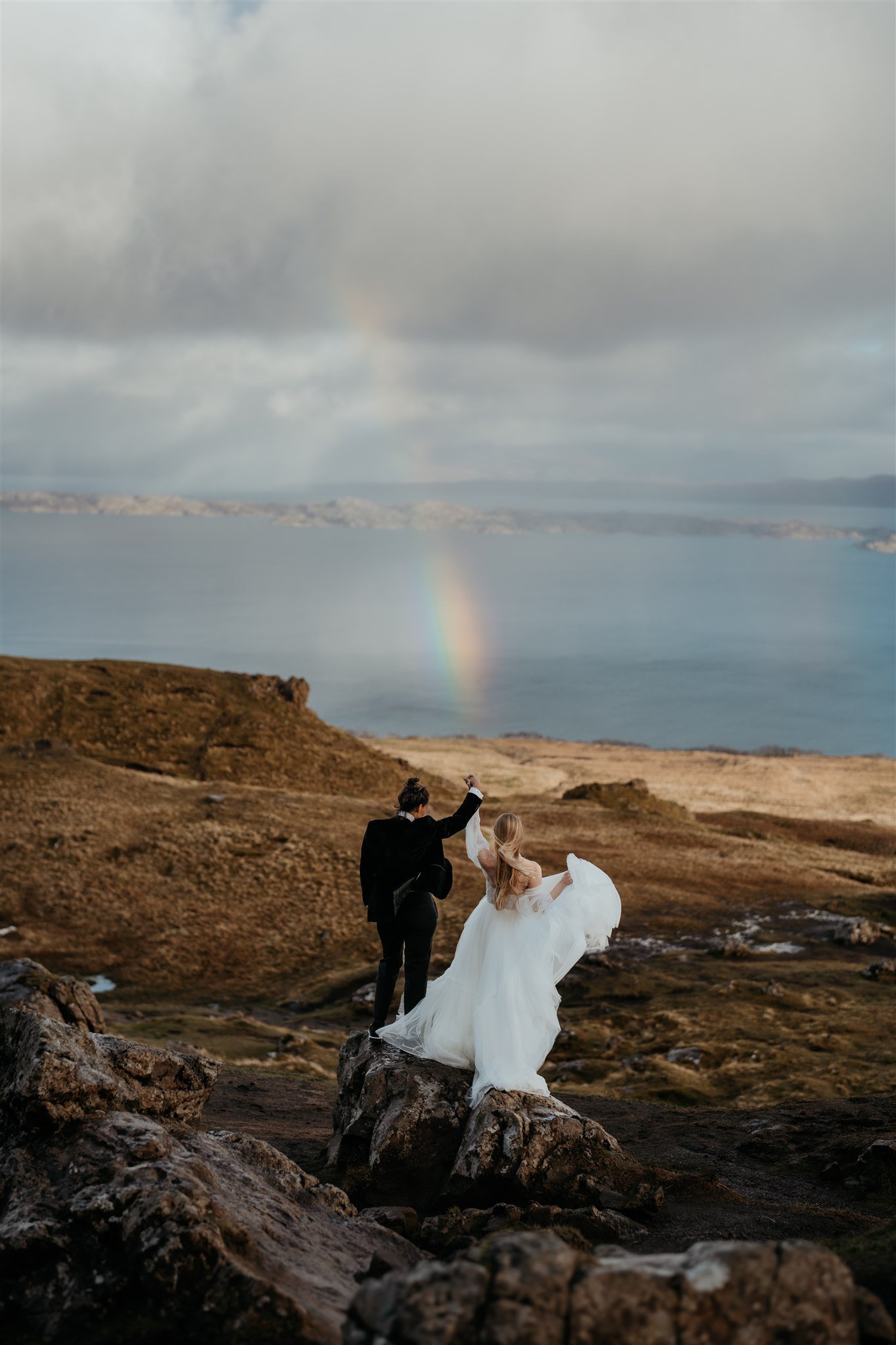 Brides cheer with a rainbow shining in the background at their Isle of Skye elopement