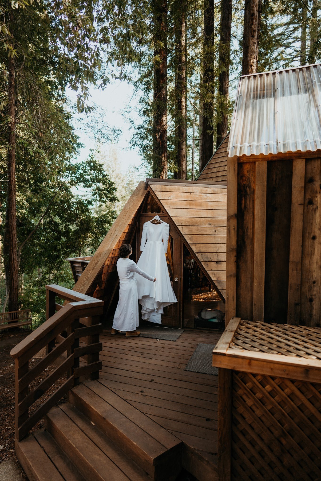 Bride adjusting white wedding dress hanging from A-Frame cabin door at Sequoia Retreat Center
