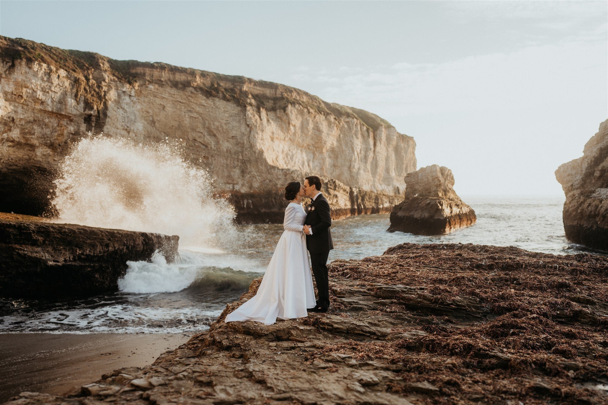 Bride and groom kiss while waves crash against the cliffs in the background