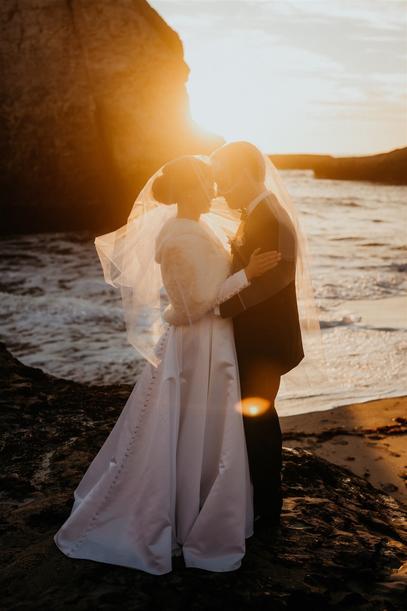 Bride and groom stand under a veil on the beach at sunset