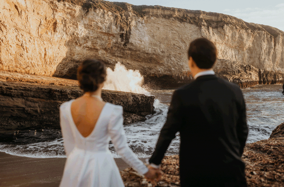 Bride and groom hold hands and smile at each other while waves crash on the shore