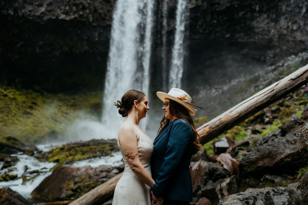 Two brides hold hands while standing in front of a waterfall at their Columbia River Gorge elopement