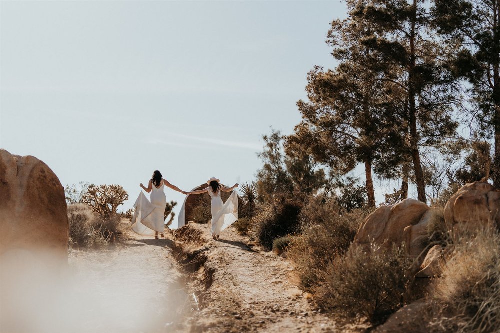 Two brides hold hands while running through the desert at their Joshua Tree elopement
