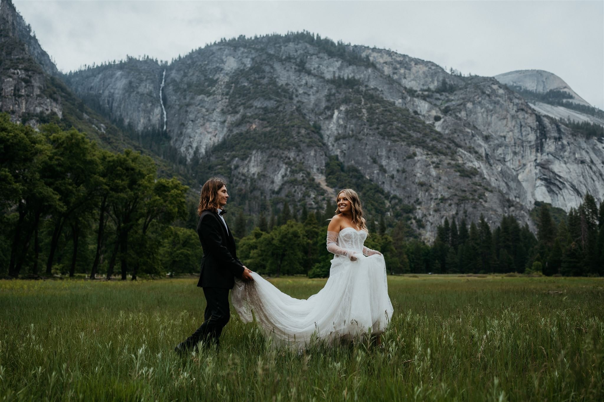 Groom holds bride's train while walking across El Capitan meadow at Yosemite elopement