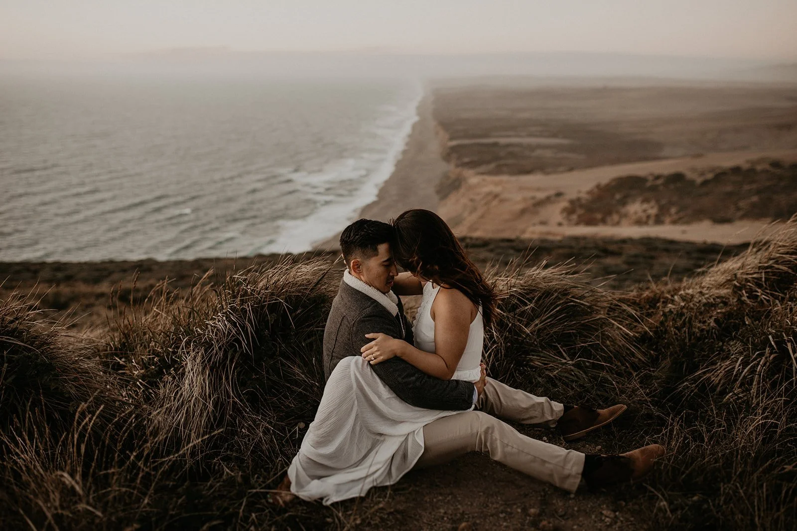 Bride and groom sit on a grassy overlook during their California engagement session at Point Reyes National Seashore. 