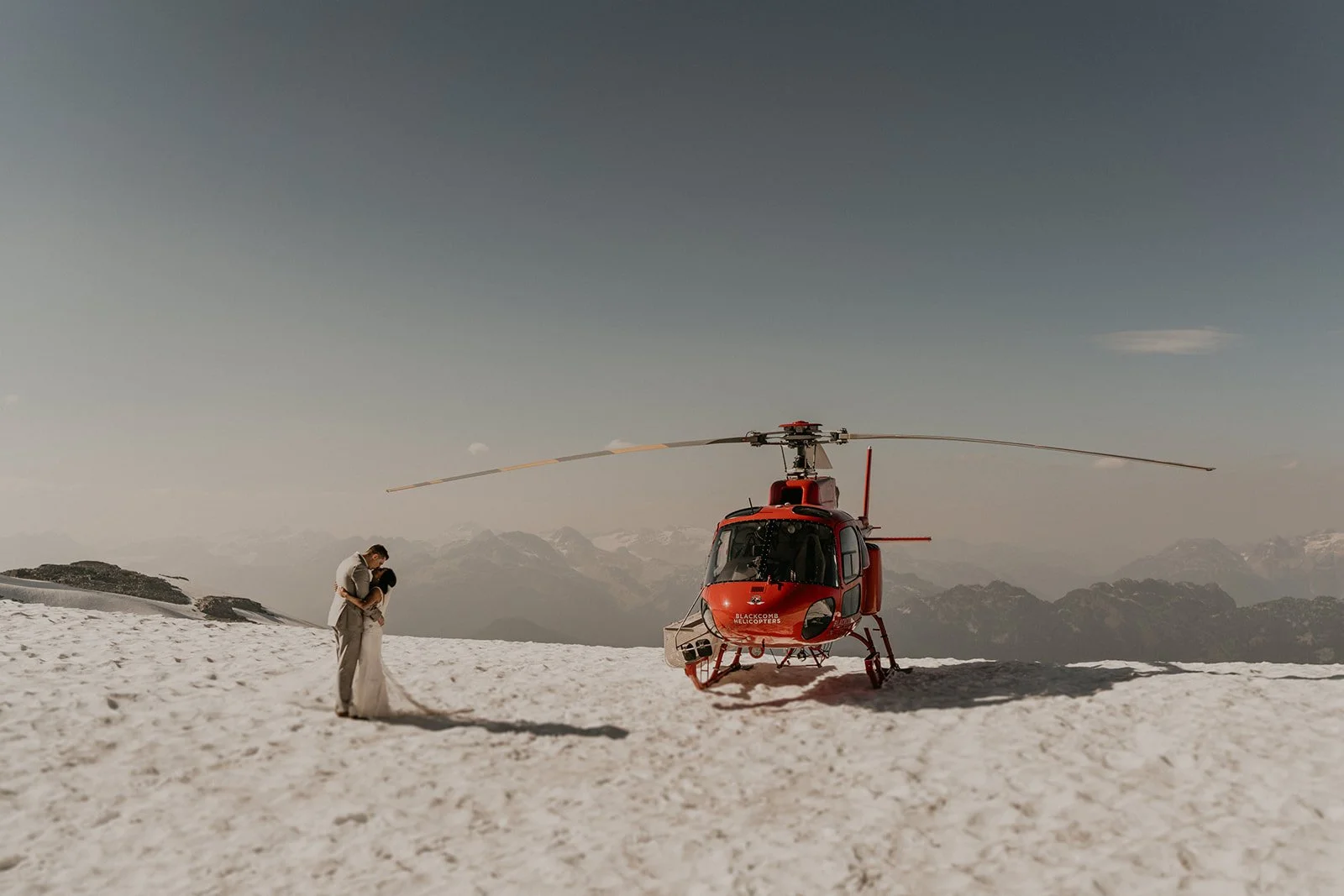 Bride and groom kissing in front of red helicopter in Whistler mountains