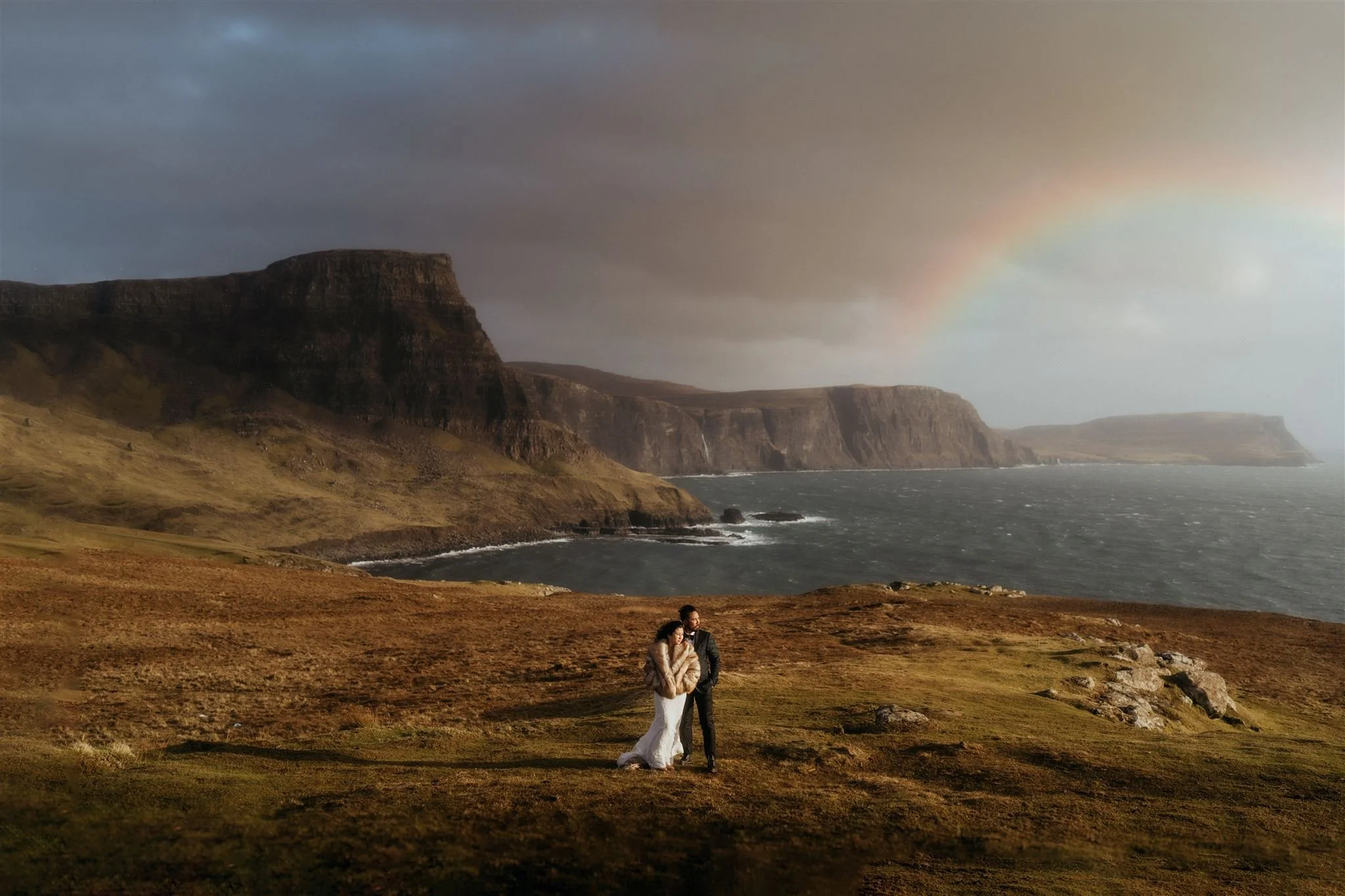 Bride and Groom standing on a field in Scotland with cliffs, ocean, and a rainbow view