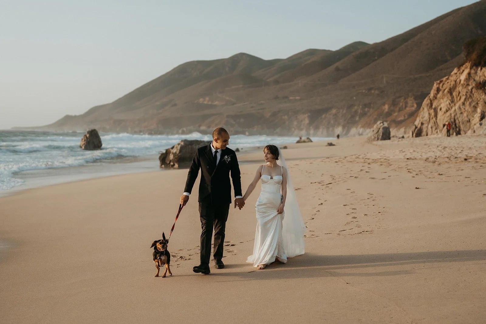 Bride and groom hold hands while walking their dog across the beach at their Big Sur elopement