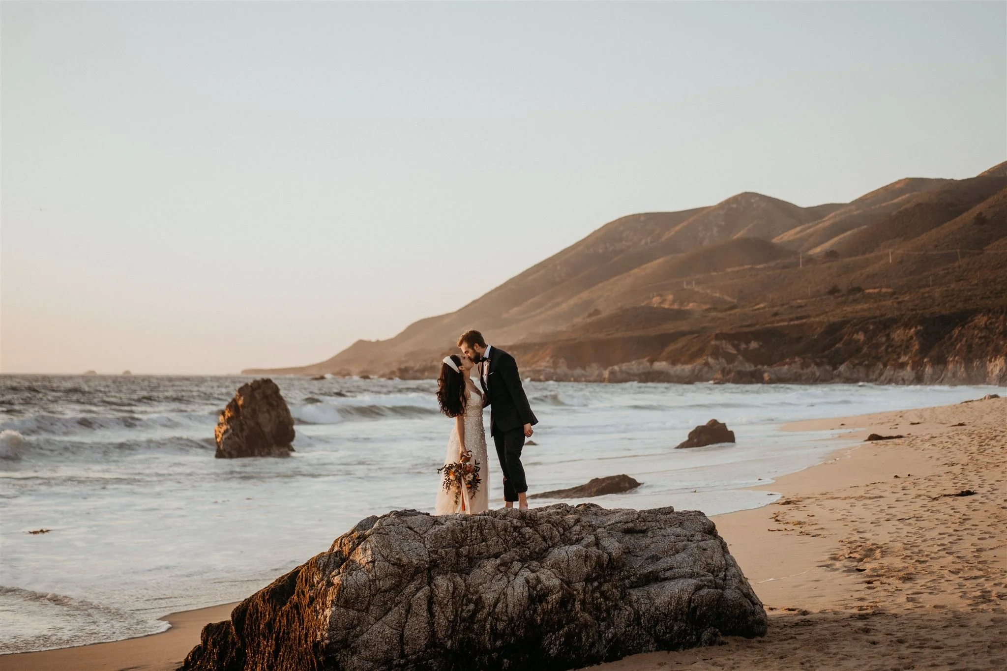 Bride and groom stand on a rock in the middle of a sandy beach during their Mendocino, California elopement. 
