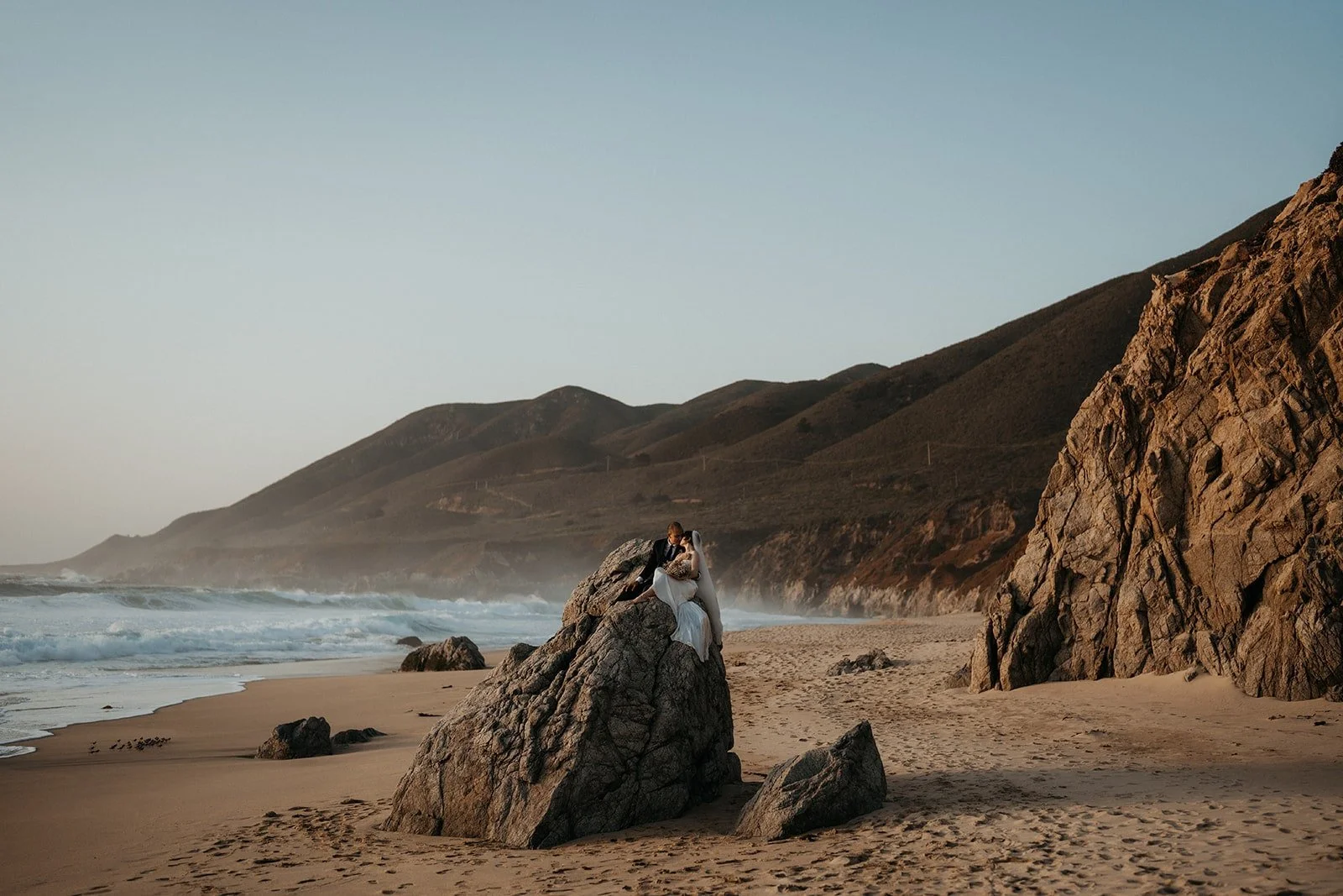 Bride and groom sit on a rock on the beach during their sunset elopement in Big Sur