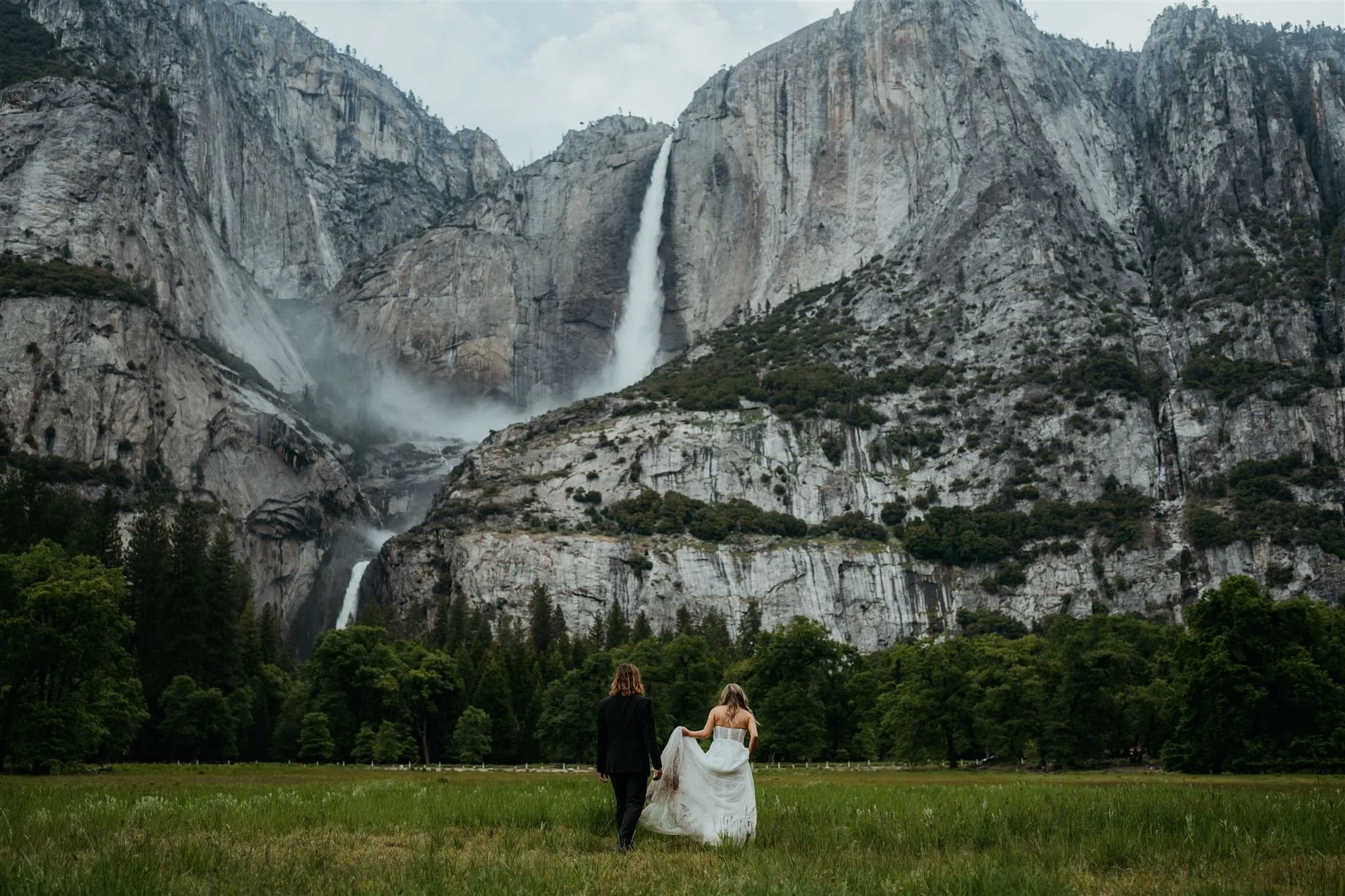 Groom holding bride's dress train while walking across the meadow toward Yosemite Falls