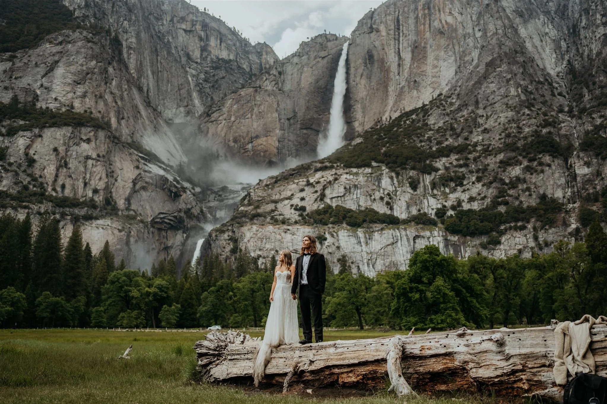 Bride and groom stand on a fallen log in front of Yosemite Falls during their elopement