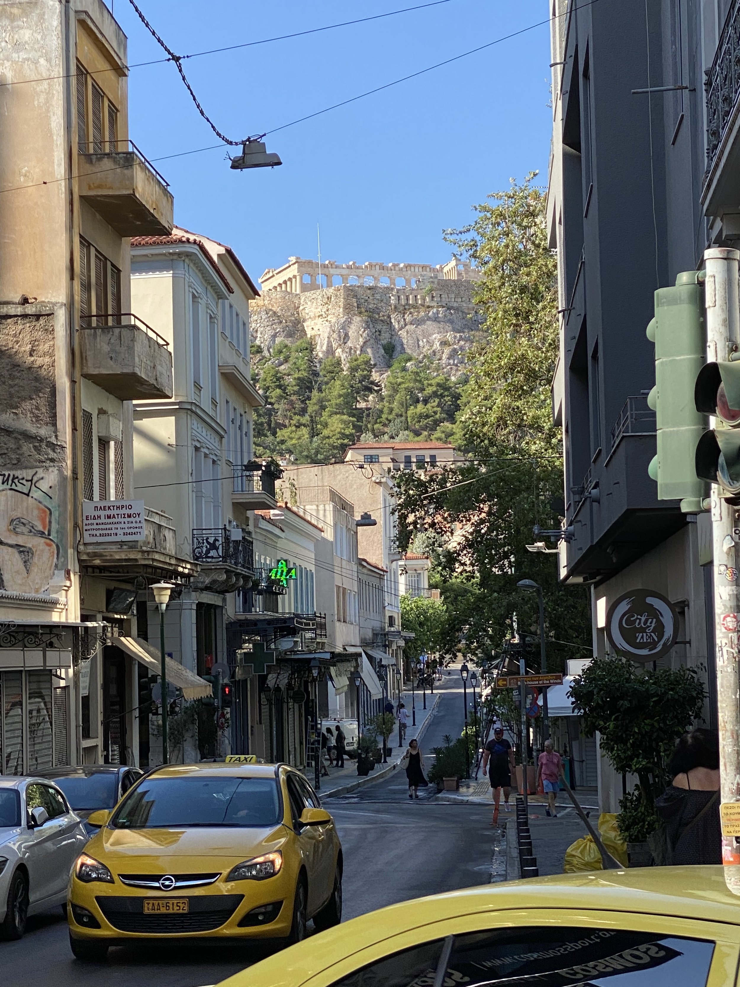 Urban street scene with cars and pedestrians, Acropolis of Athens visible in the background.