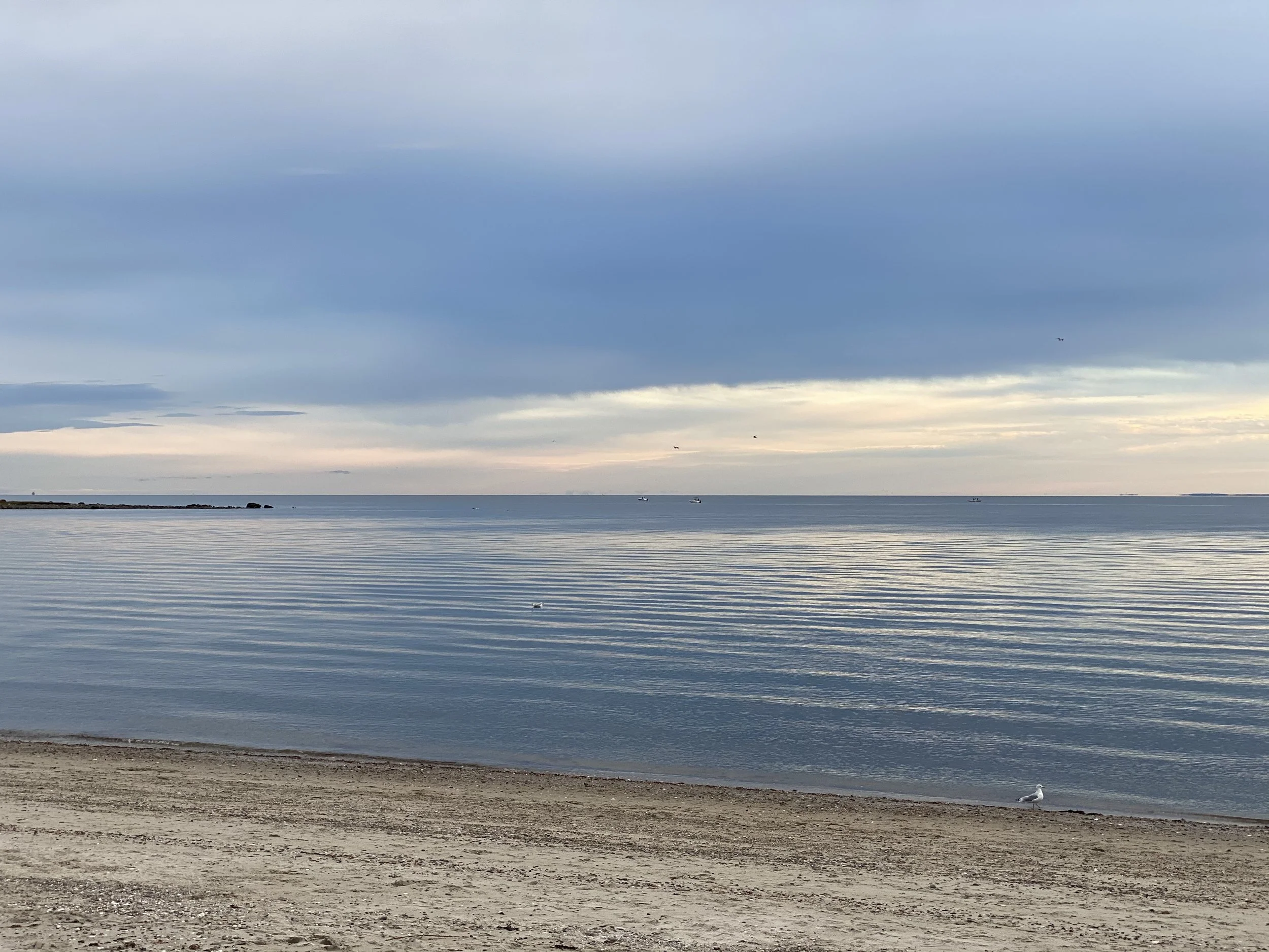Tranquil beach scene with calm sea, light waves, and sandy shore under a cloudy sky.