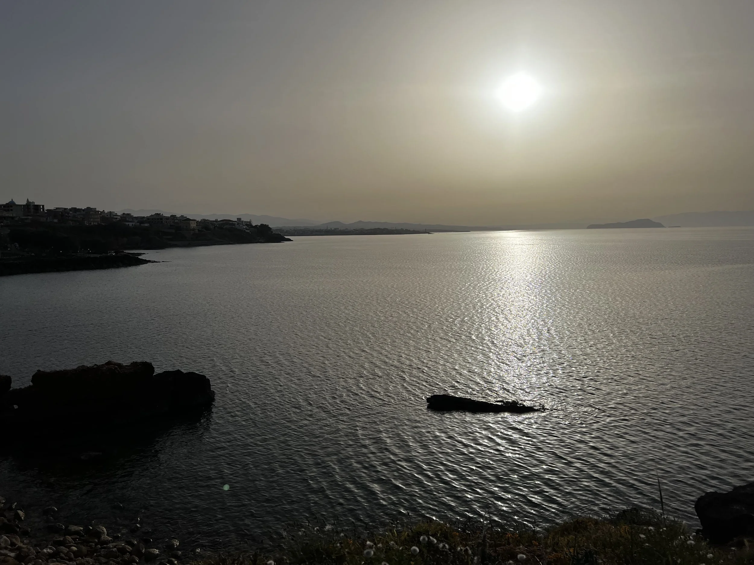 Scenic view of a calm sea with the sun setting silhouetting distant hills and coastal buildings