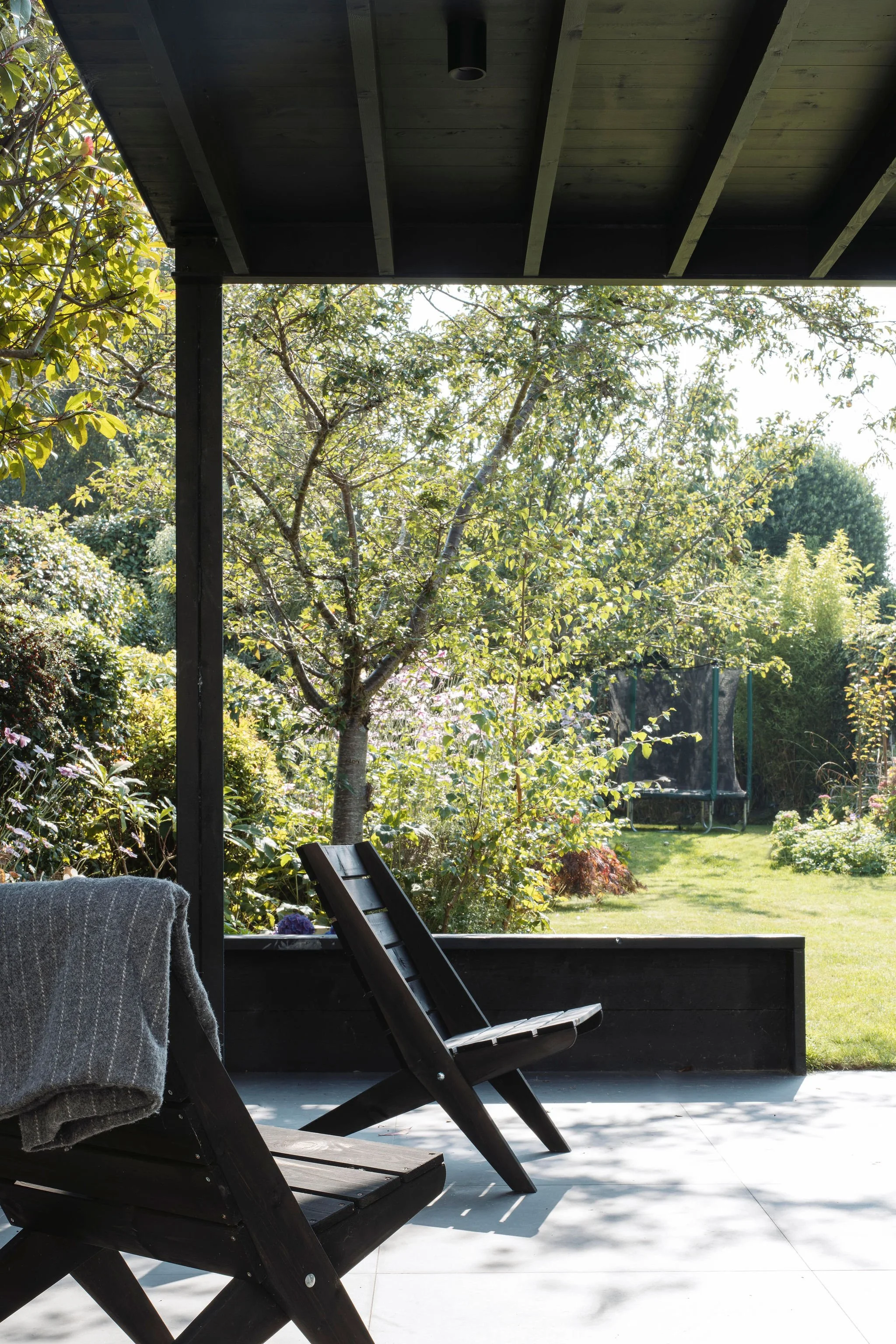 A covered patio with two black wooden chairs facing a lush garden with trees and bushes, sunlight filtering through the foliage.