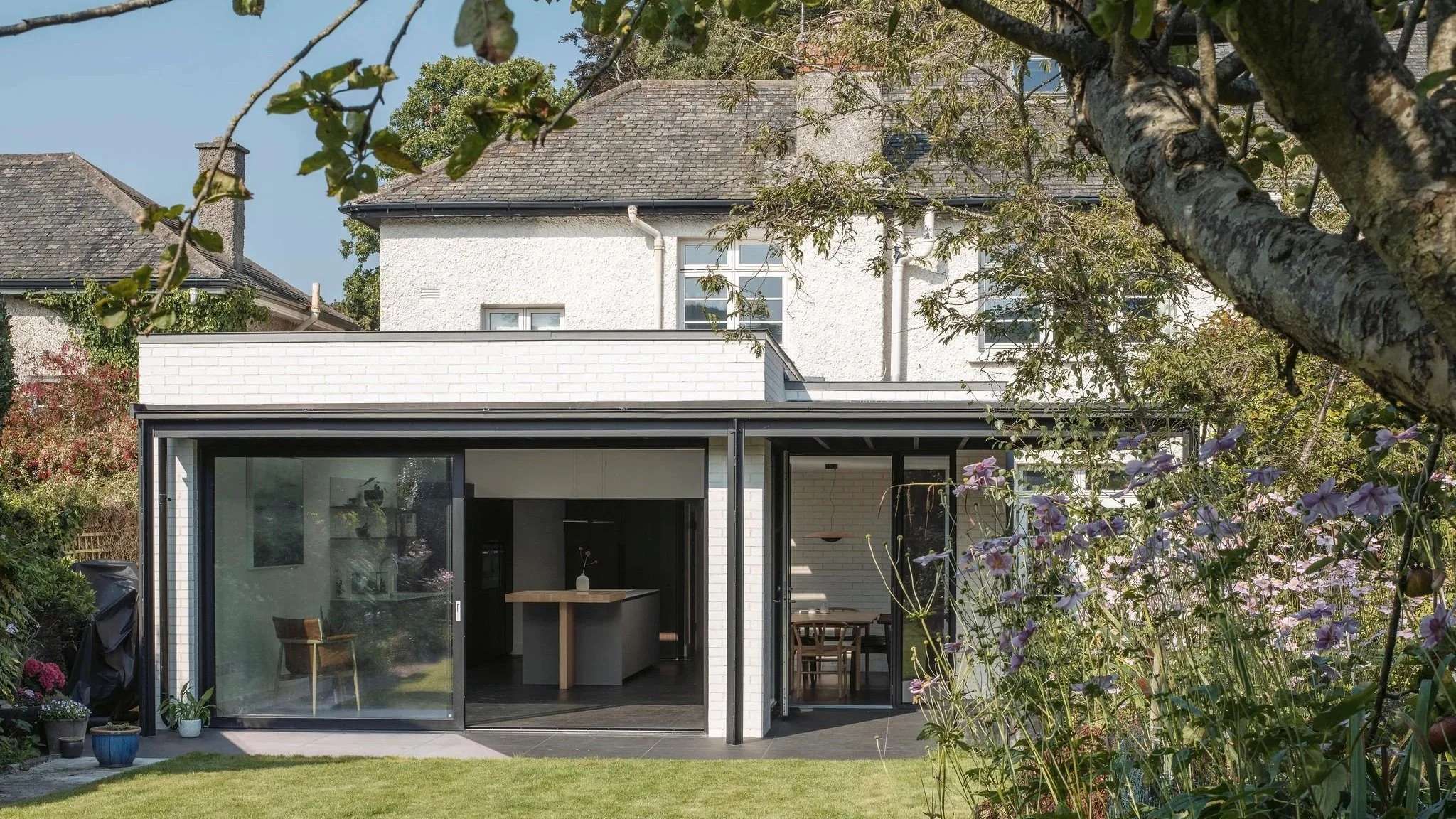 Modern house with a garden, featuring large glass sliding doors, a lawn, and trees and flowers in the foreground.