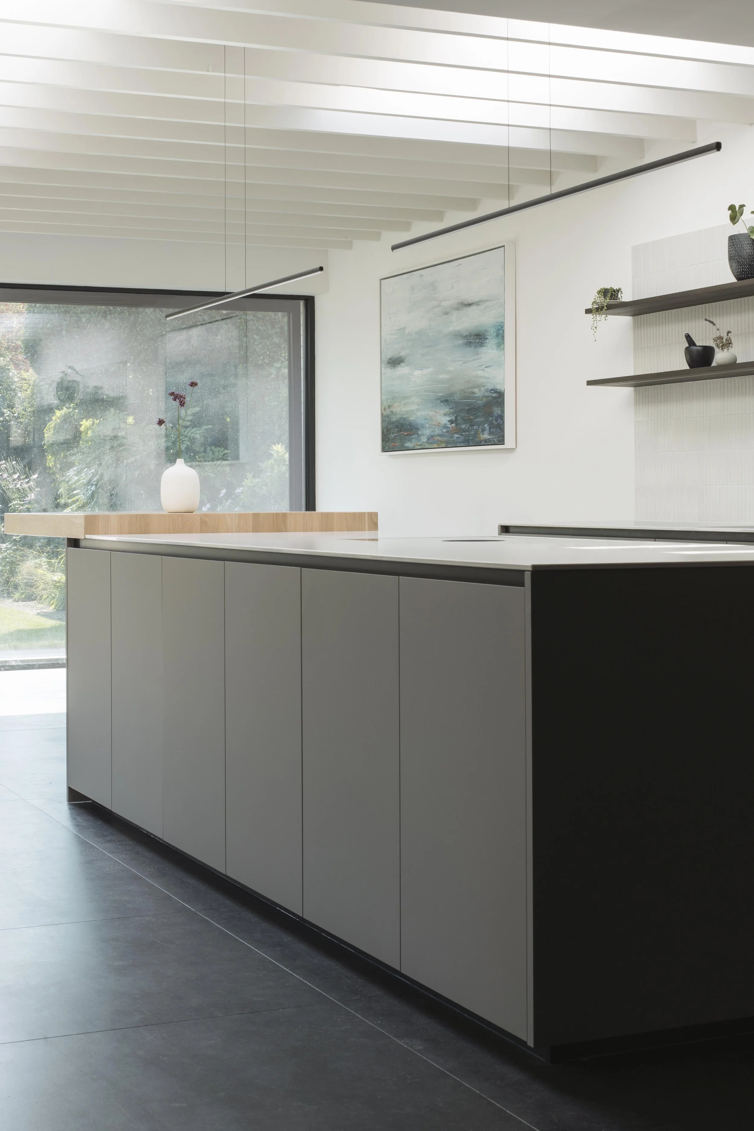 Modern kitchen with grey cabinets, black countertop, large window with view of greenery, abstract wall art, and floating shelves with small decorative items.