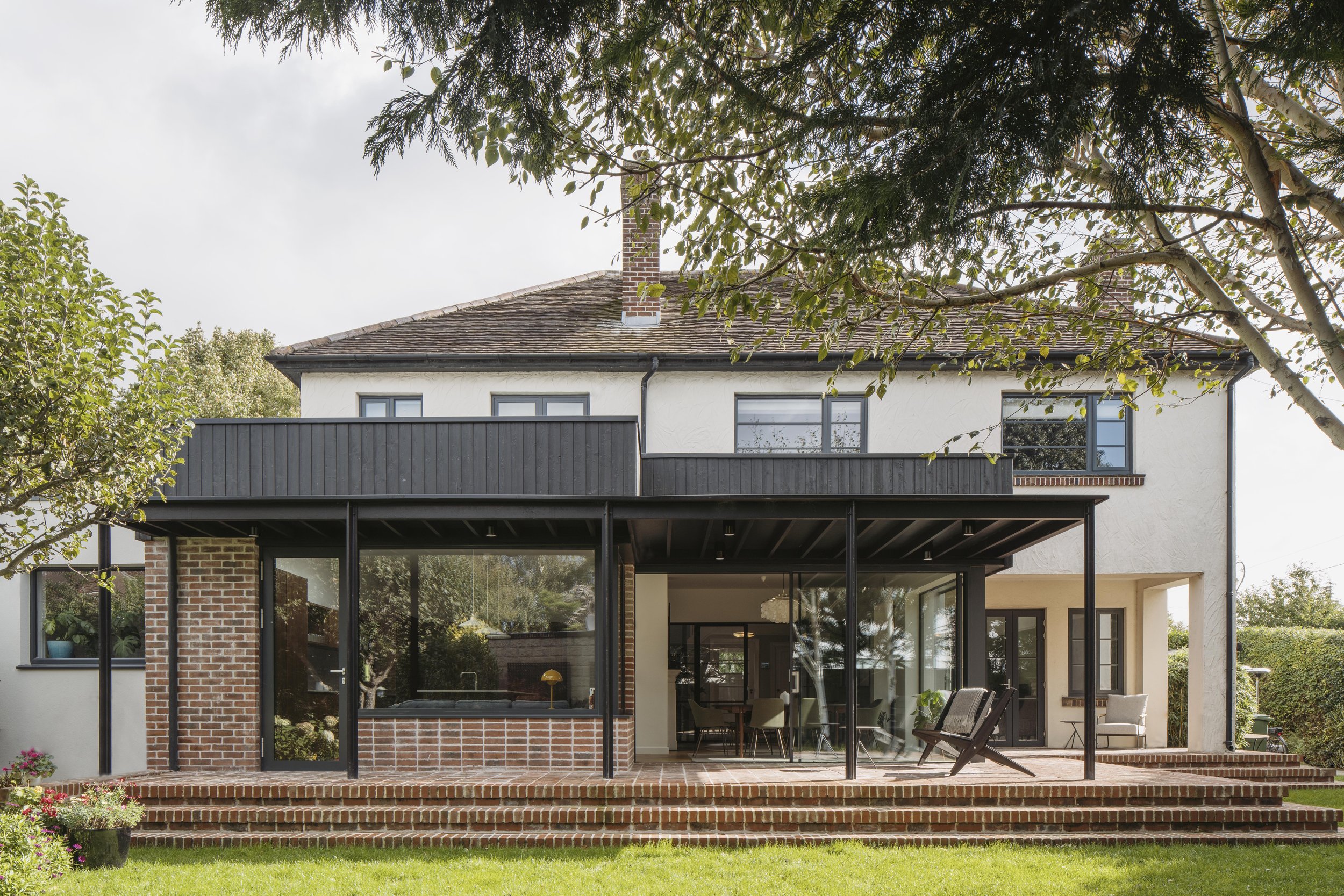Modern two-story house with a white exterior, black window frames, a brick chimney, and an extensive black balcony. The backyard features a brick patio, outdoor seating, and lush green grass with trees.
