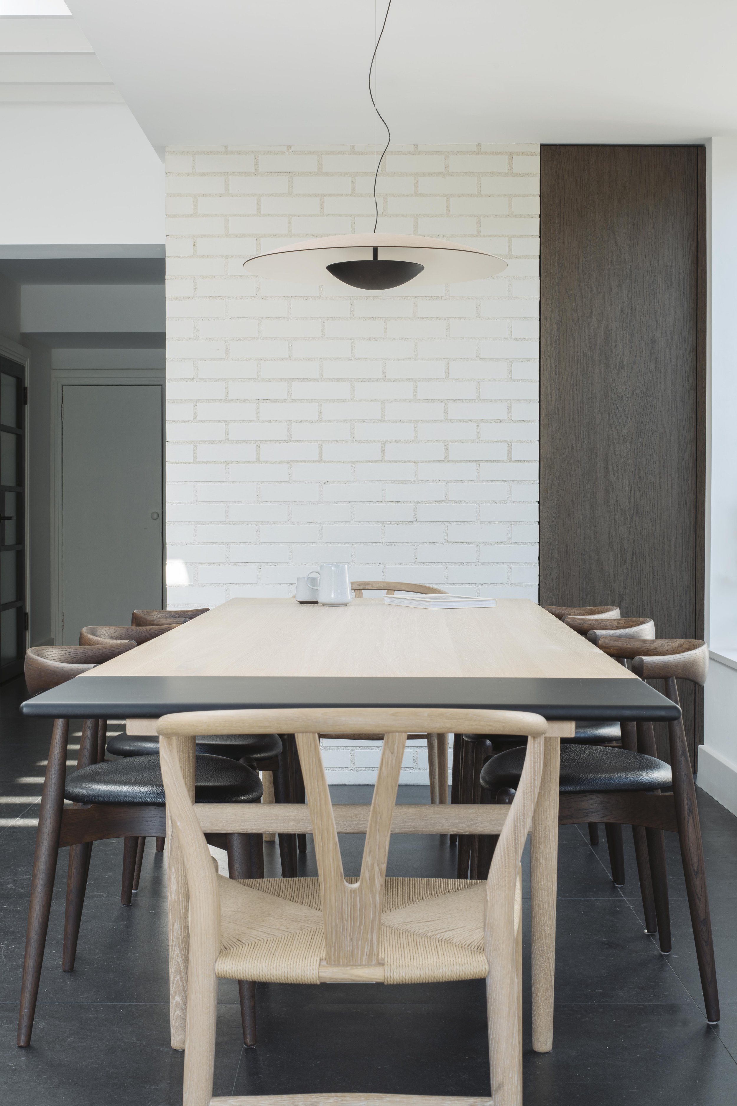 Minimalist dining room with a large wooden table surrounded by mix of wooden chairs, white brick wall, modern pendant light fixture hanging from ceiling, and dark wood paneling on wall.