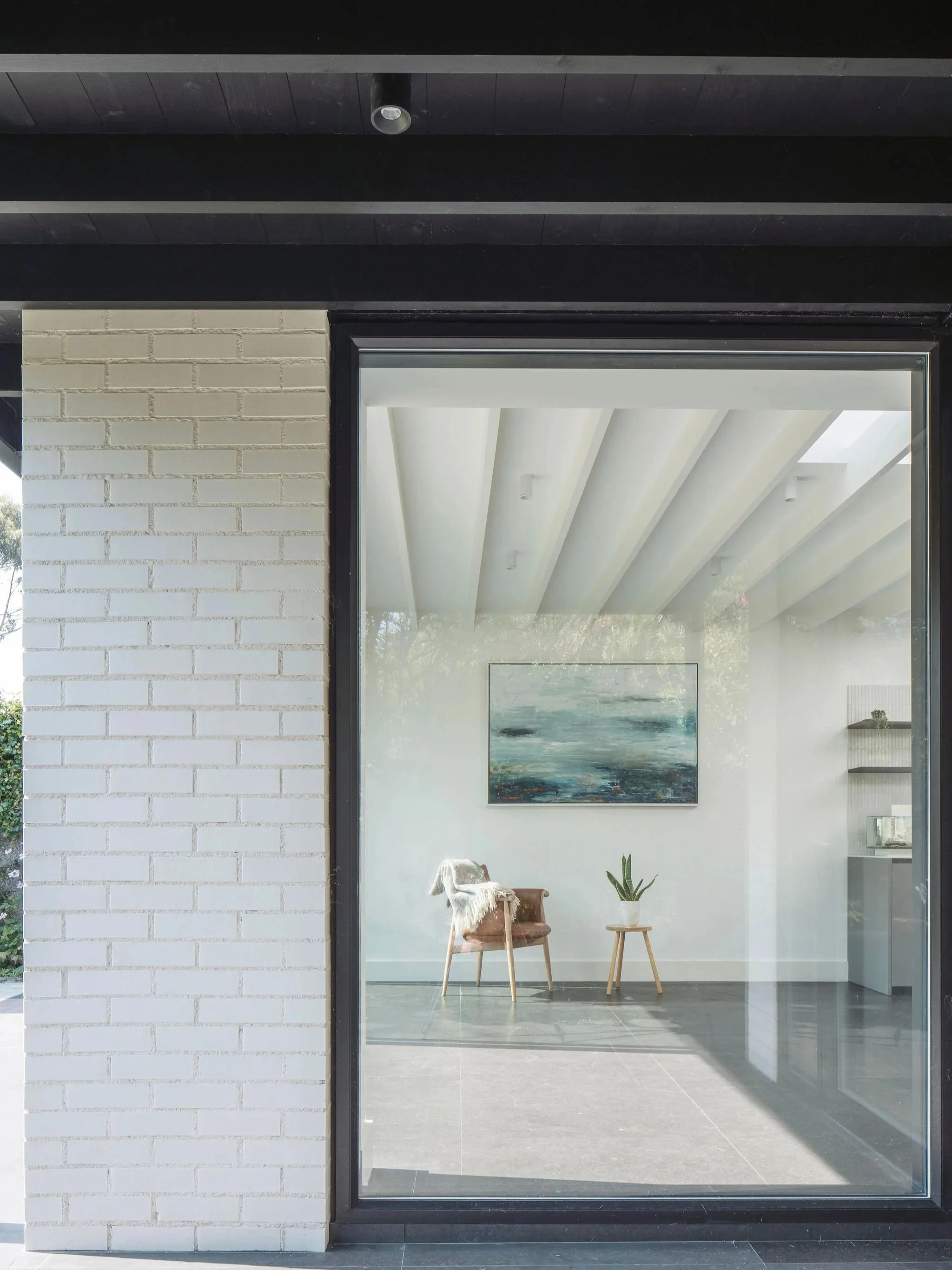 View through a large glass window into a modern, minimalist interior with a wooden chair, a potted plant on a small wooden table, and a landscape painting on the white wall.