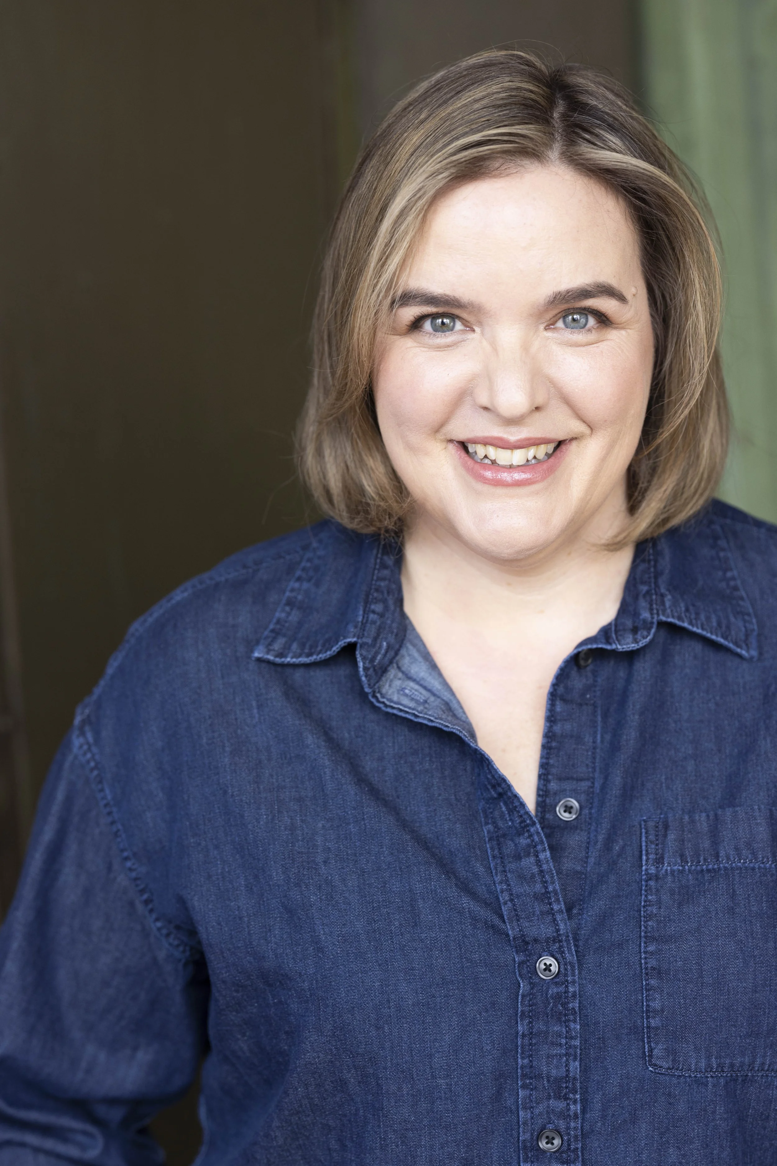 Close-up of a middle-aged woman with shoulder-length light brown hair, blue eyes, and a friendly smile, wearing a dark denim shirt. She is standing indoors with a neutral background.