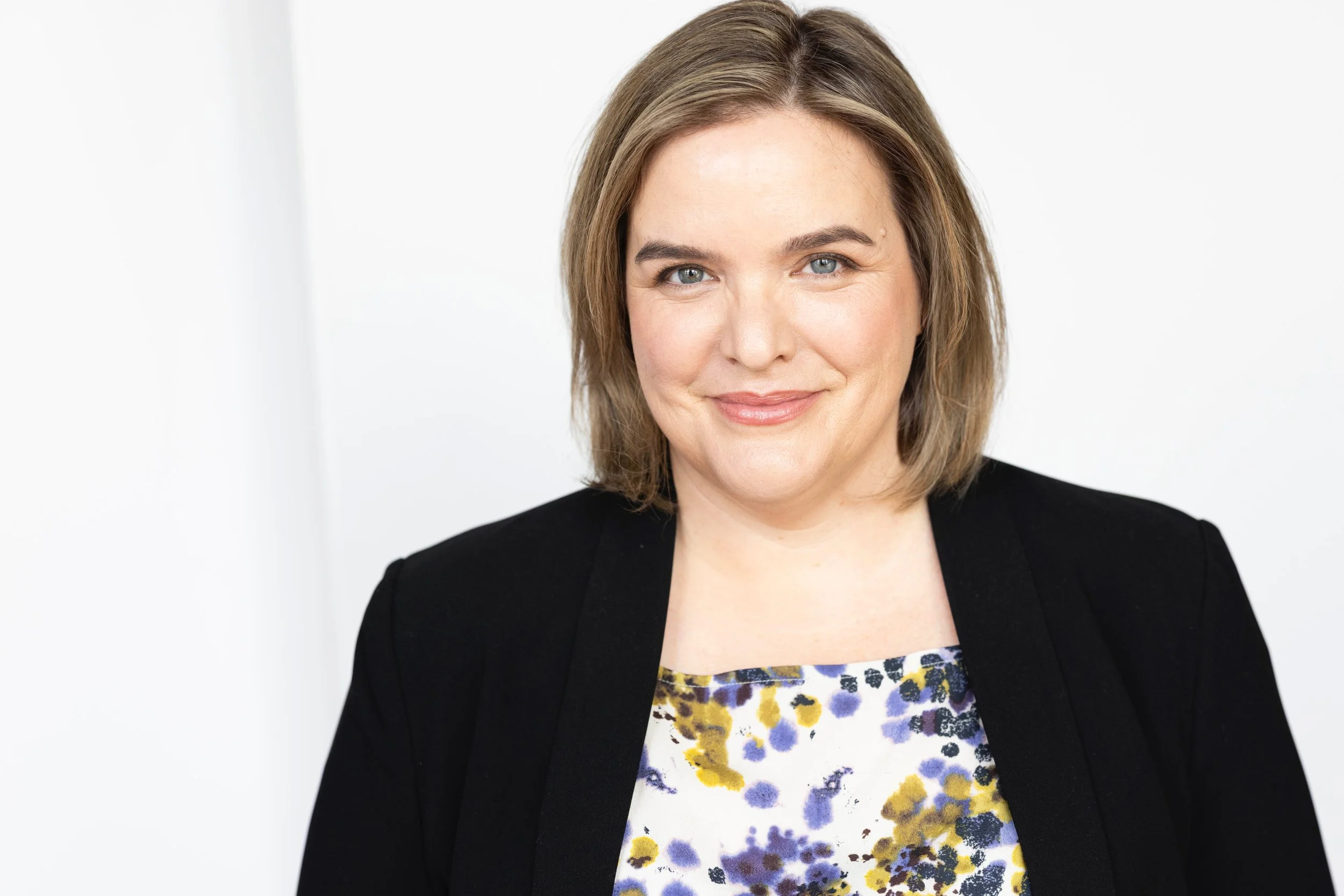 A woman with light brown hair and blue eyes dressed in a black blazer and a floral blouse, smiling against a plain white background.