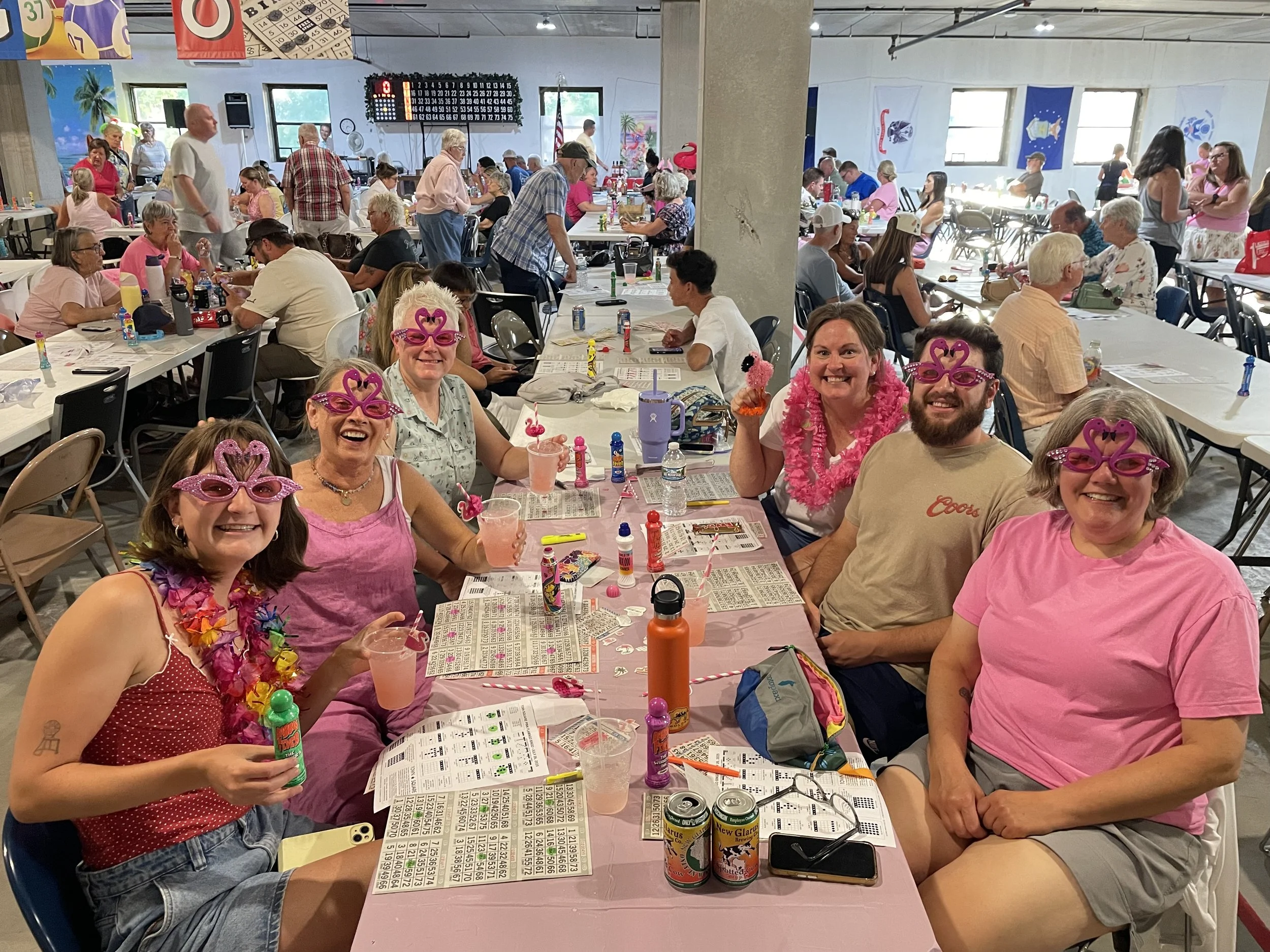 Five Bingo players wearing pink flamingo glasses and accessories smile around a table covered with Bingo cards and drinks.