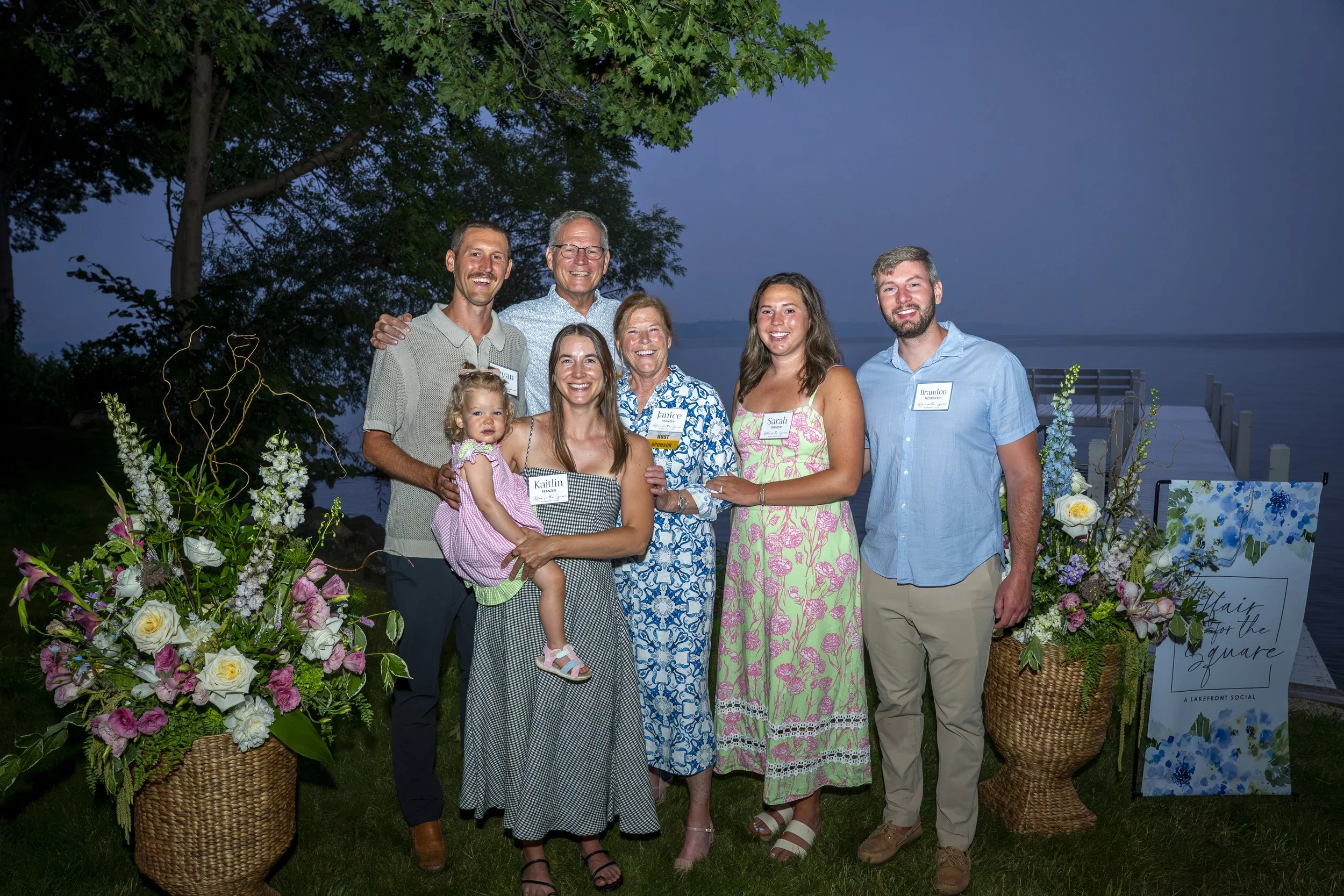 Six adults and a young child pose between colorful floral arrangements beside Green Lake during Affair for the Square.