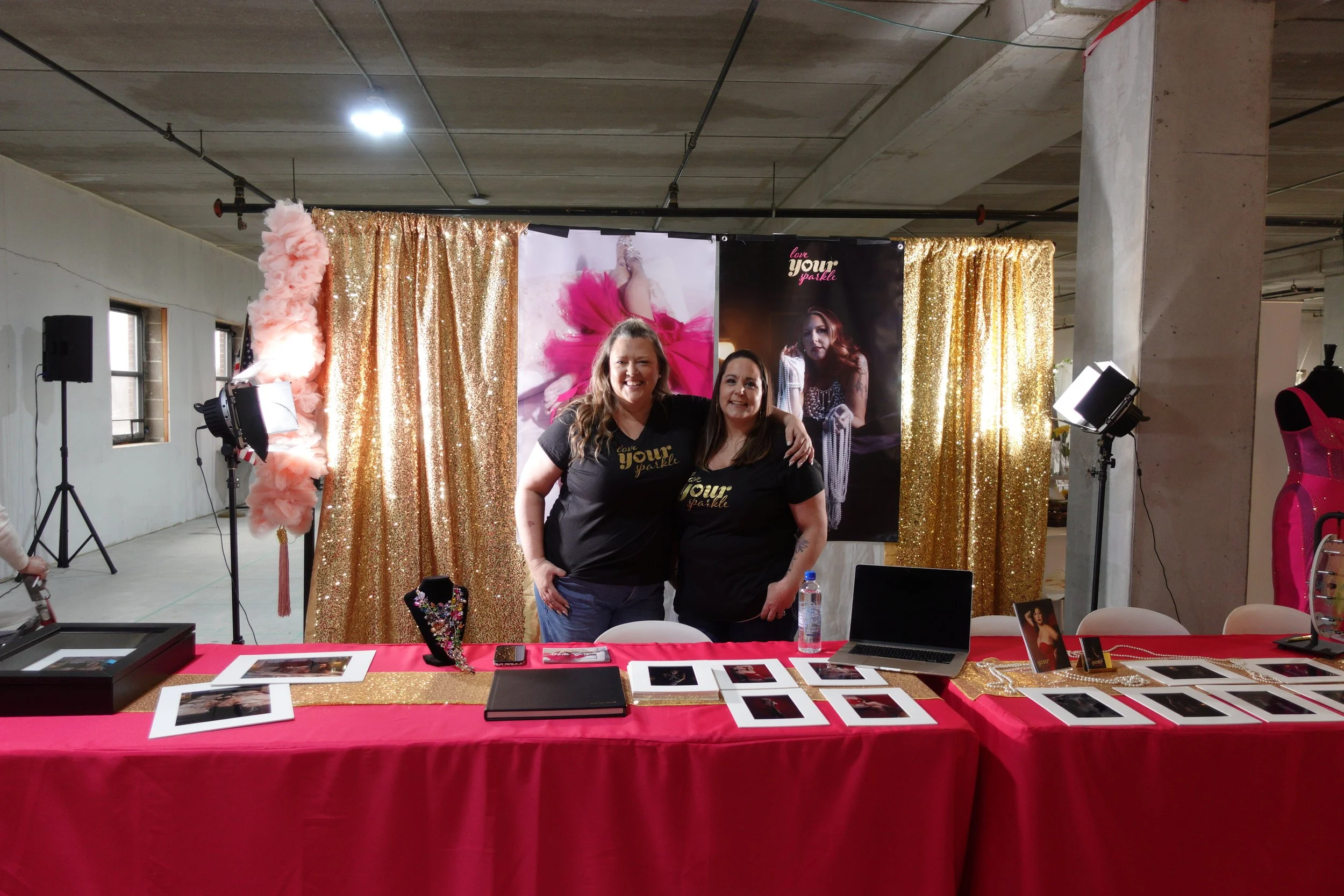 Two exhibitors pose in front of a gold sequin backdrop with photography displays and colorful decorations at the Wedding & Event Expo.