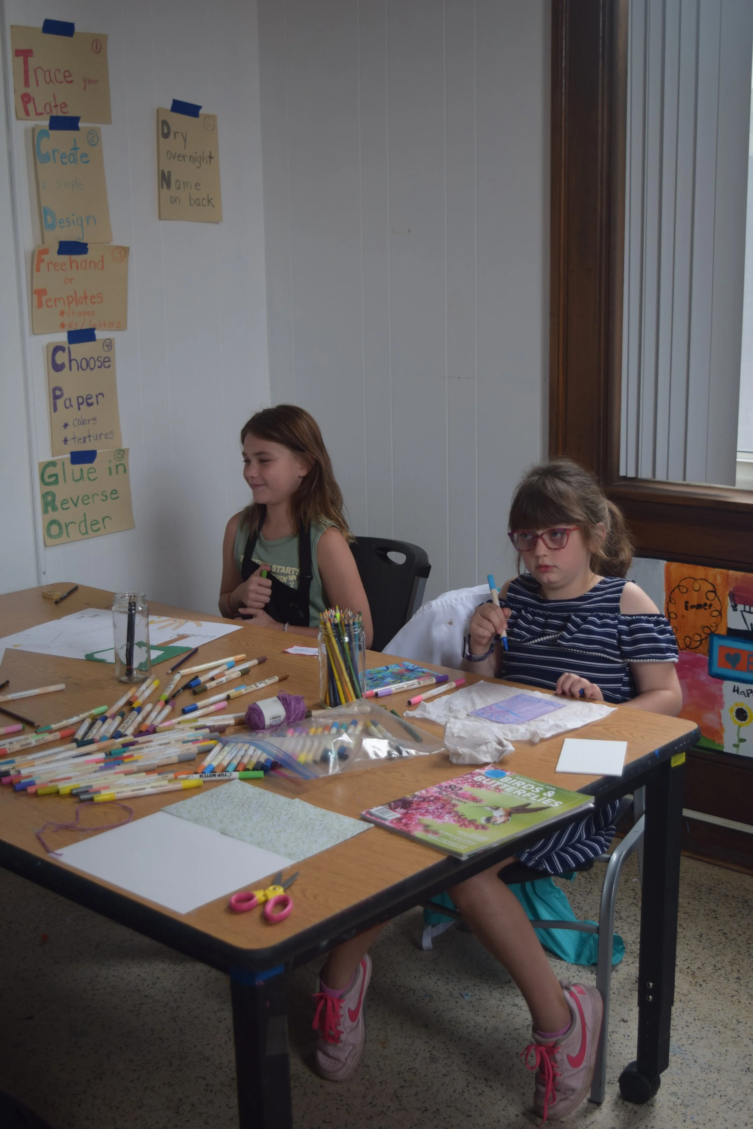 Two girls work on creative projects surrounded by markers, pencils, paper, and other craft supplies in the Town Square MakerSpace.