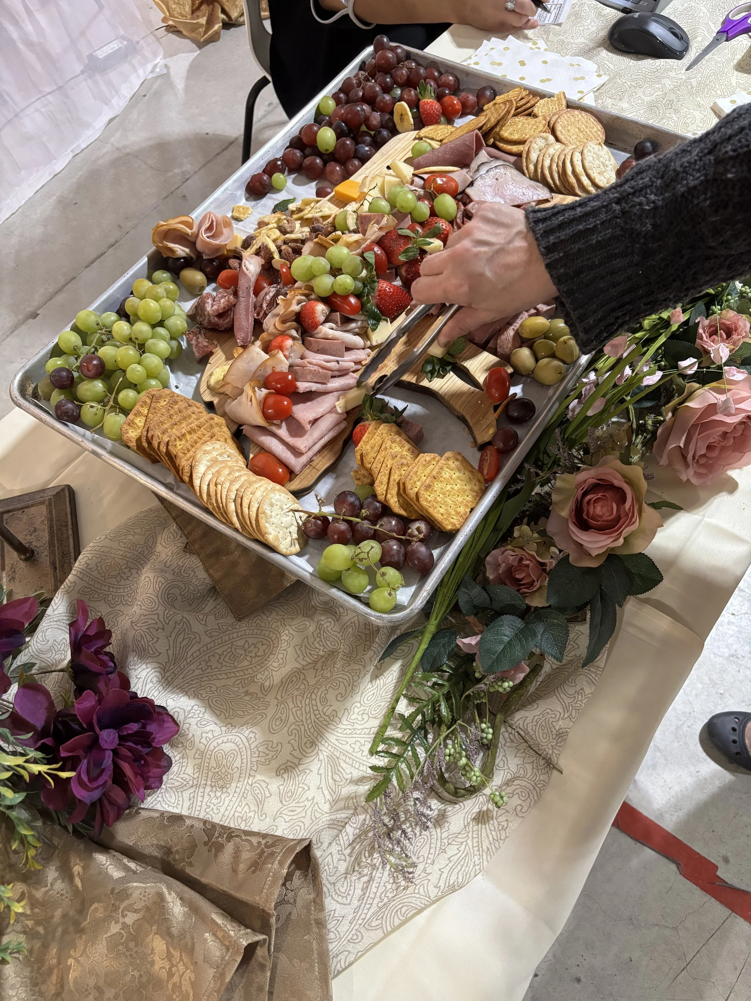 Charcuterie display featuring meats, cheeses, crackers, grapes and strawberries alongside floral arrangements at the Wedding & Event Expo.