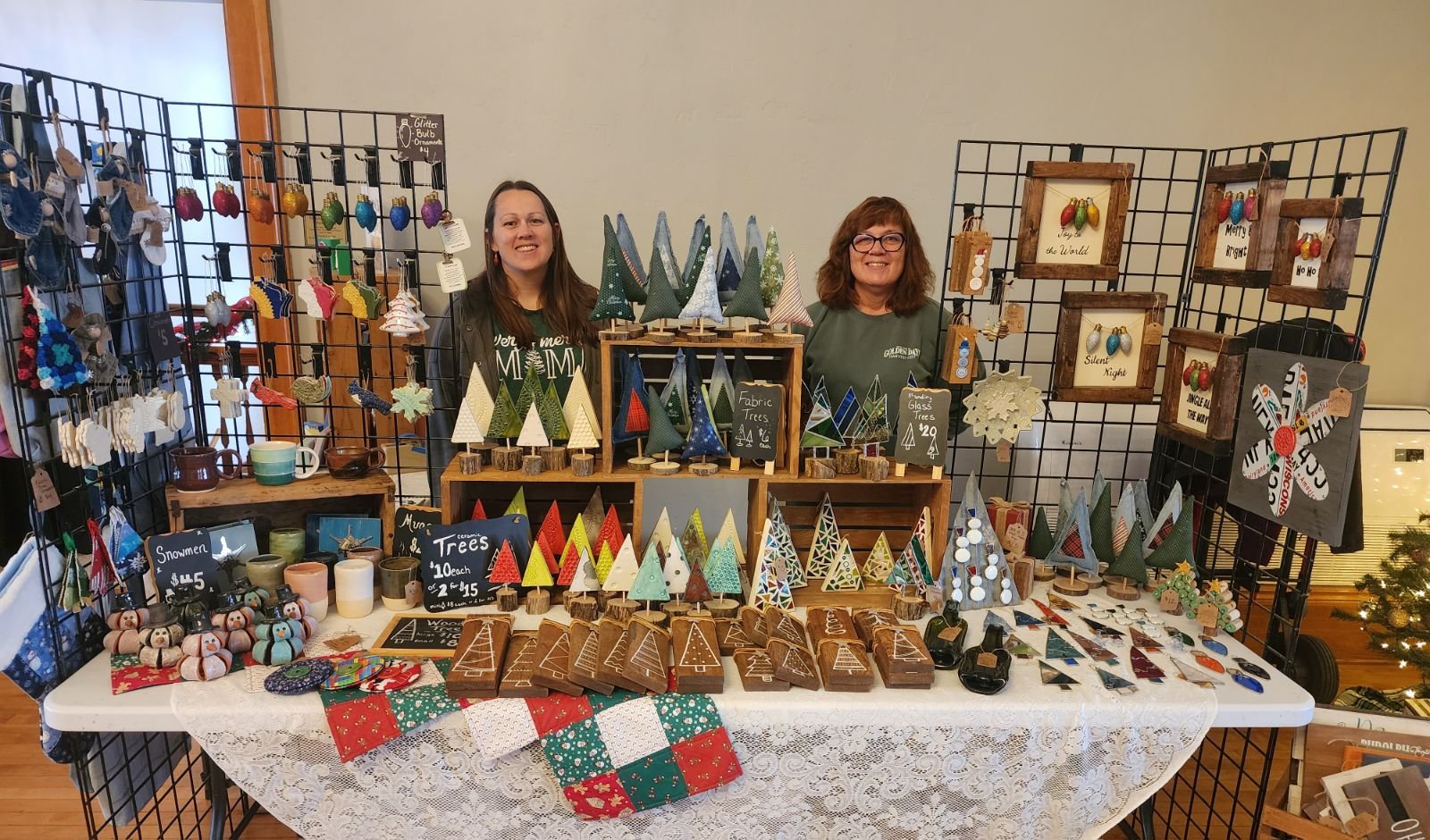 Local makers stand behind a Holiday Market booth filled with handmade trees, ornaments, pottery and festive gifts.