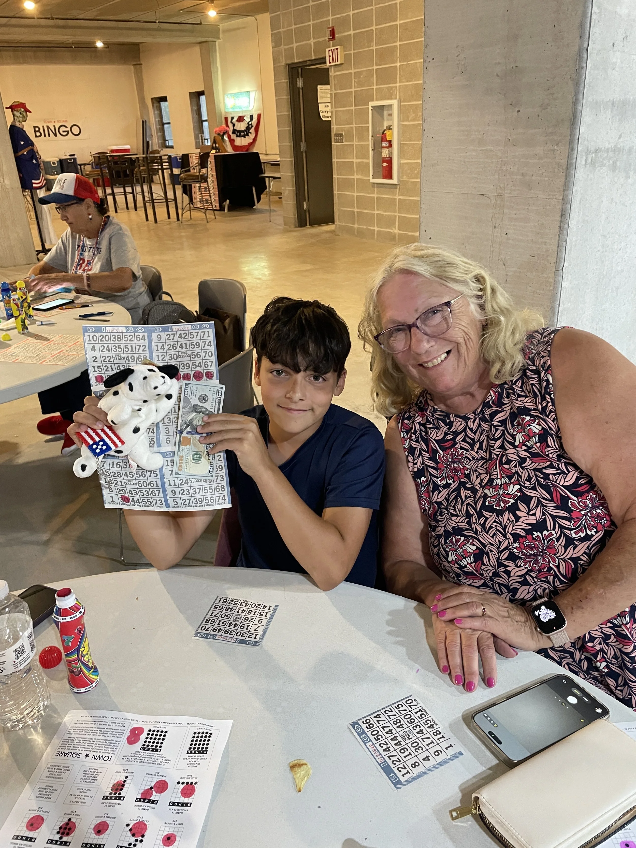 Smiling woman and young boy sitting together at a Bingo table while the boy holds Bingo cards, cash and a stuffed animal.