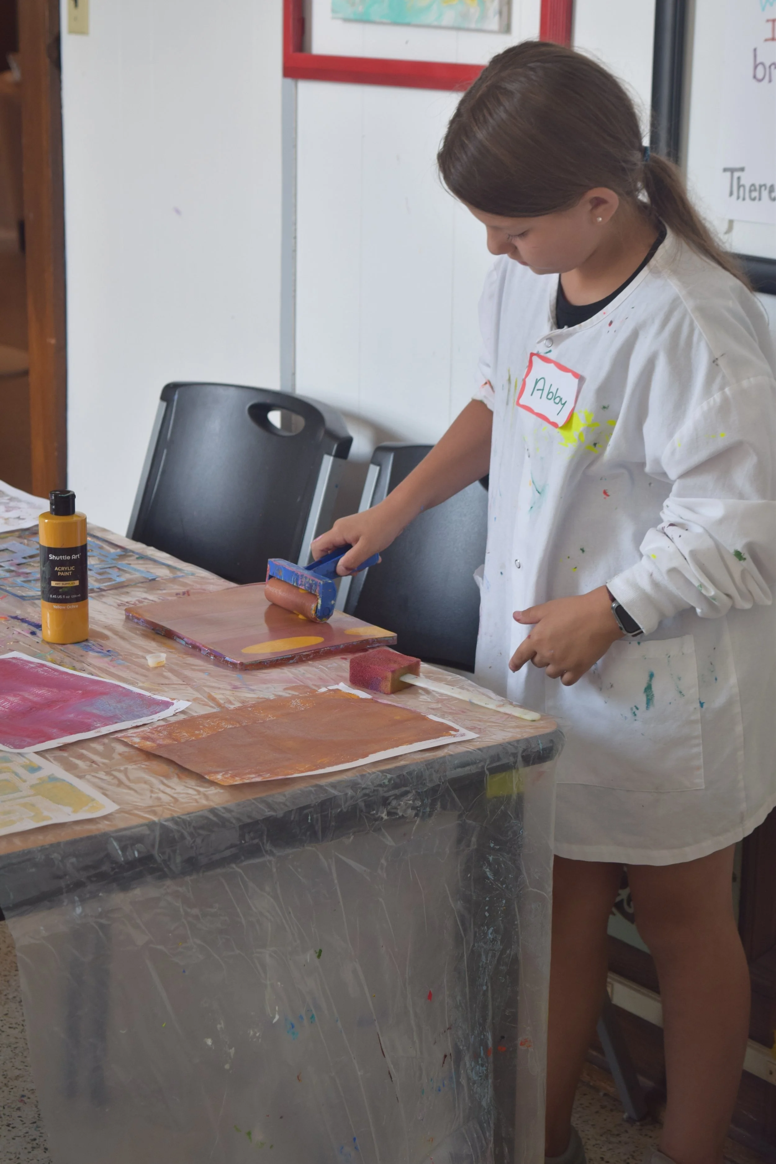 Girl uses a small roller to apply paint to an art project during a creative activity in the Town Square MakerSpace.