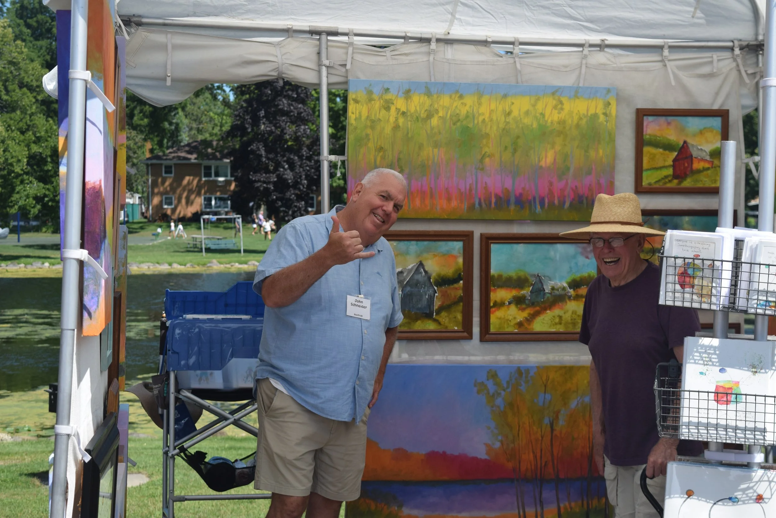 Two men pose inside an outdoor art booth surrounded by colorful landscape paintings at the Green Lake Fine Art Show.