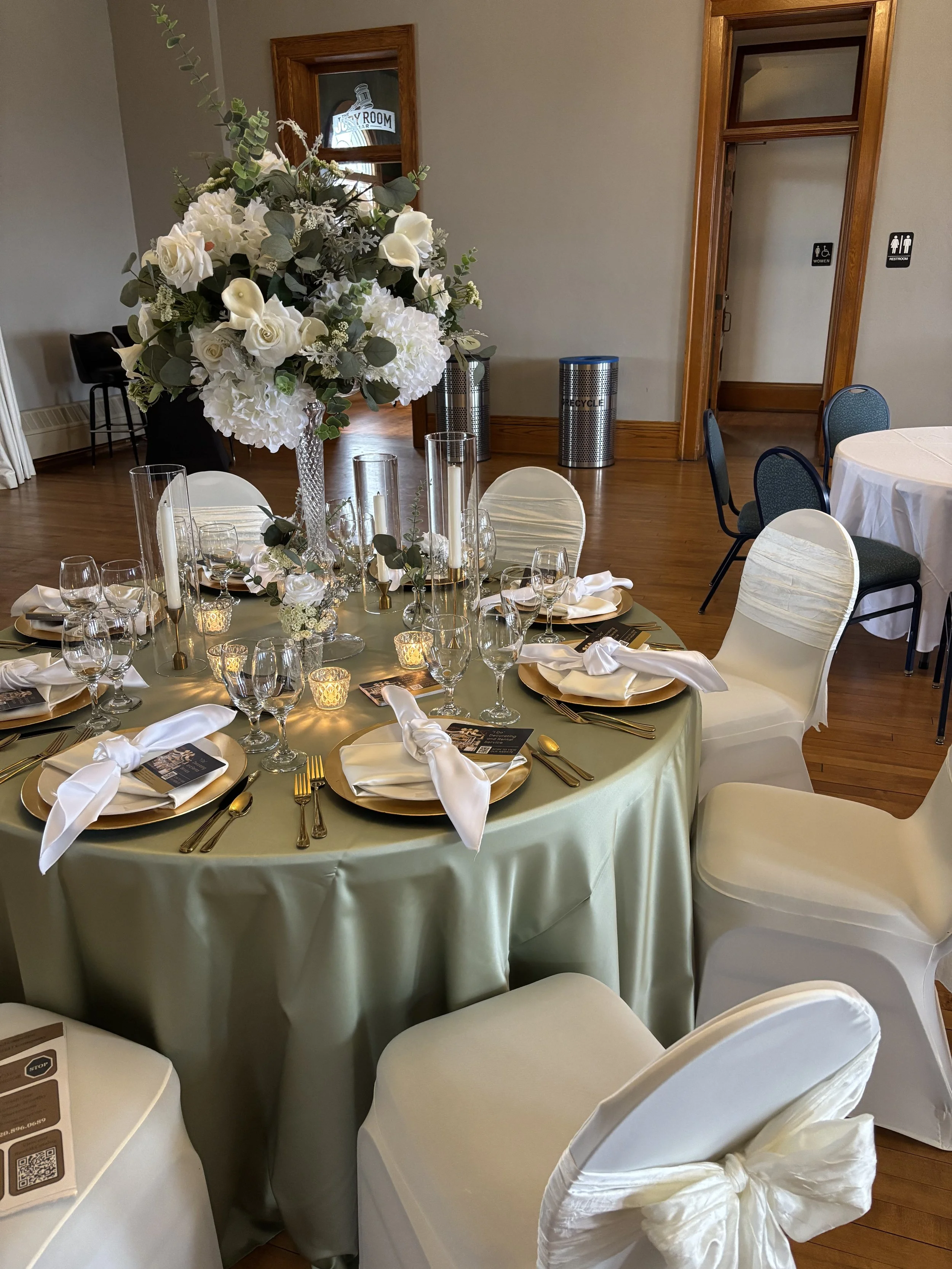 Elegantly decorated reception table with white floral centerpieces, sage green linens, gold place settings and candles inside Town Square Community Center.