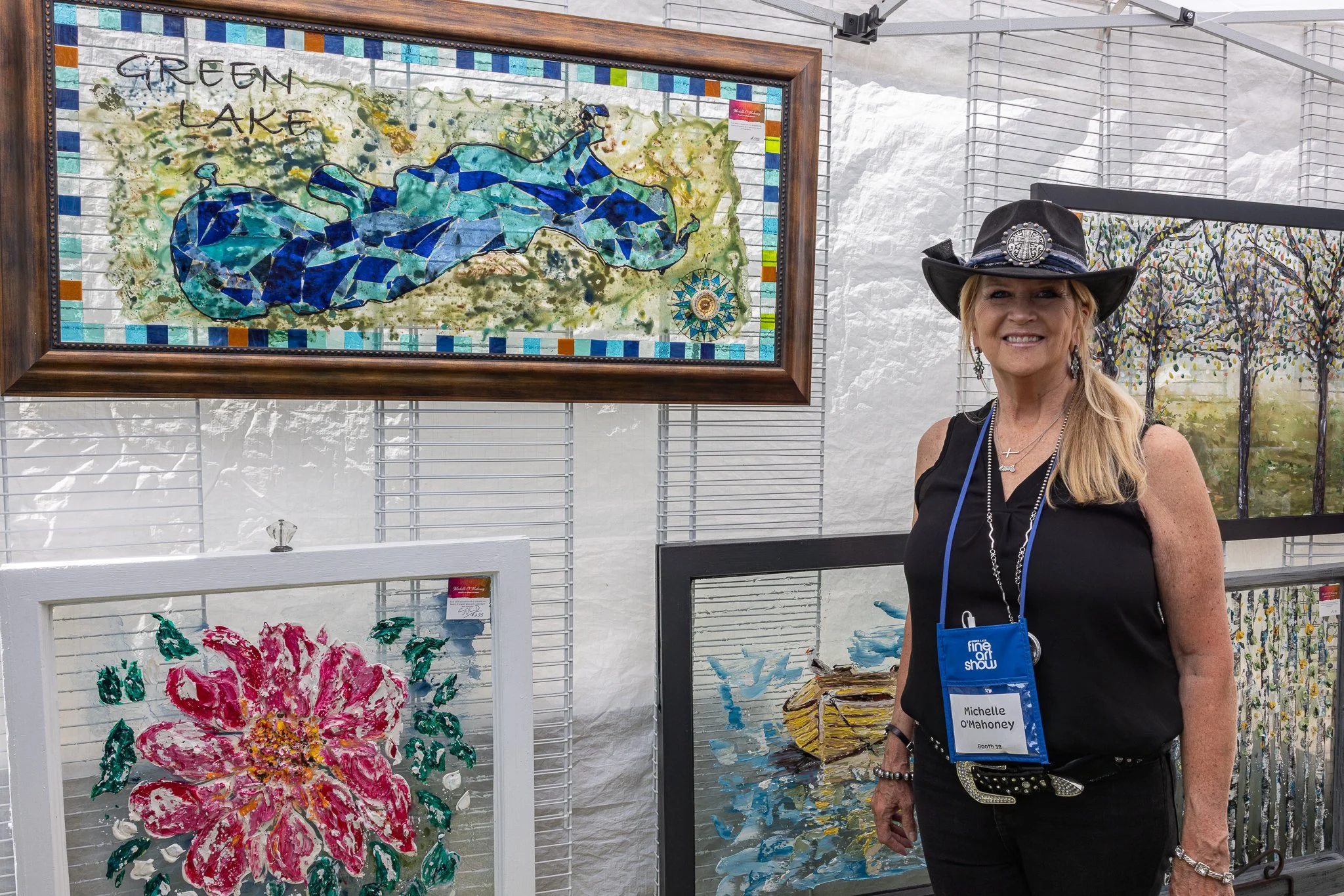Artist Michelle O'Mahoney stands beside her paintings and a framed Green Lake-inspired artwork at the Green Lake Fine Art Show.