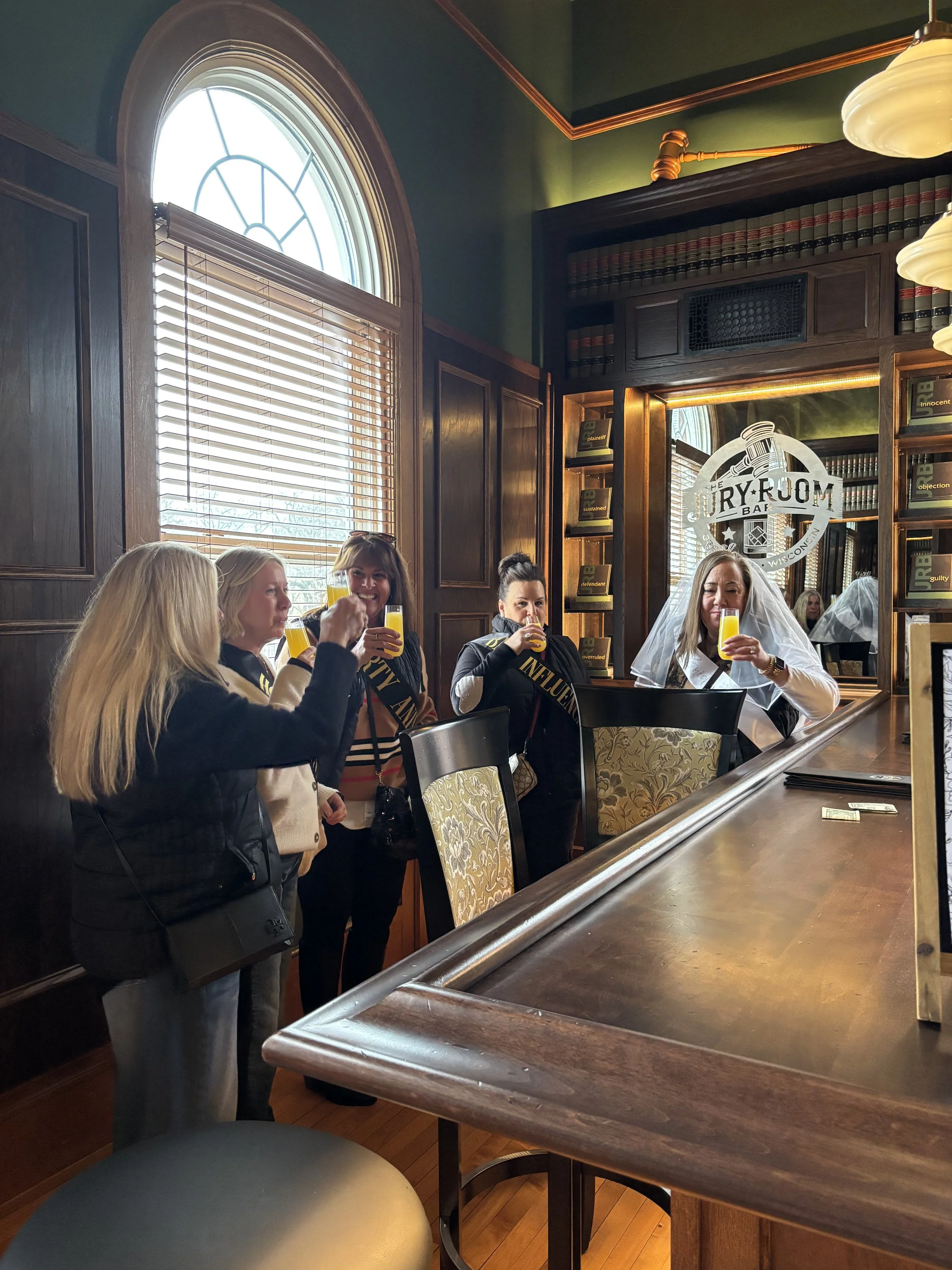 Group of women, including a bride wearing a white veil, raise drinks together inside the historic Jury Room Bar at Town Square Community Center.