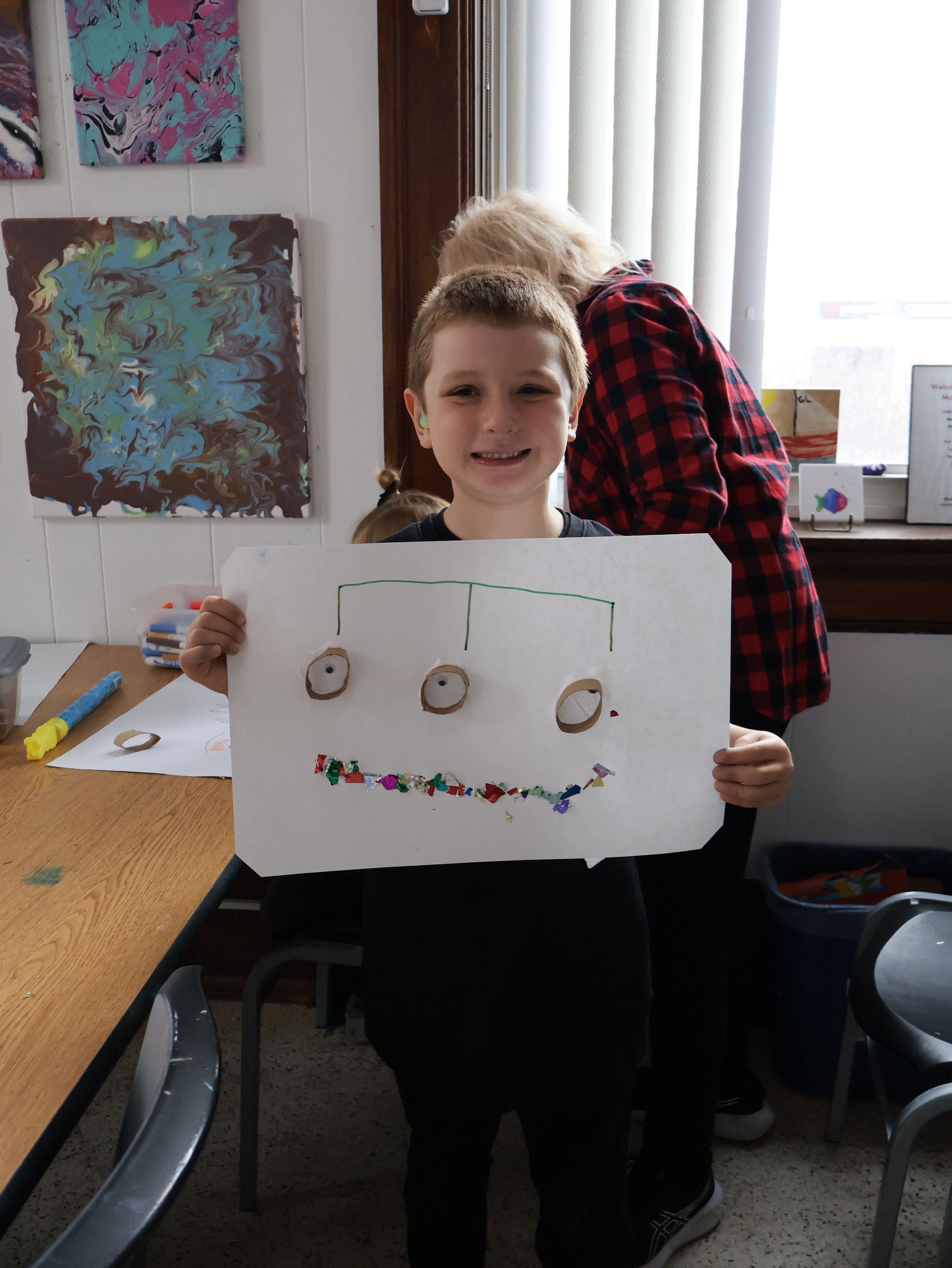 Smiling boy holds up his finished mixed-media art project during a kids activity in the MakerSpace.