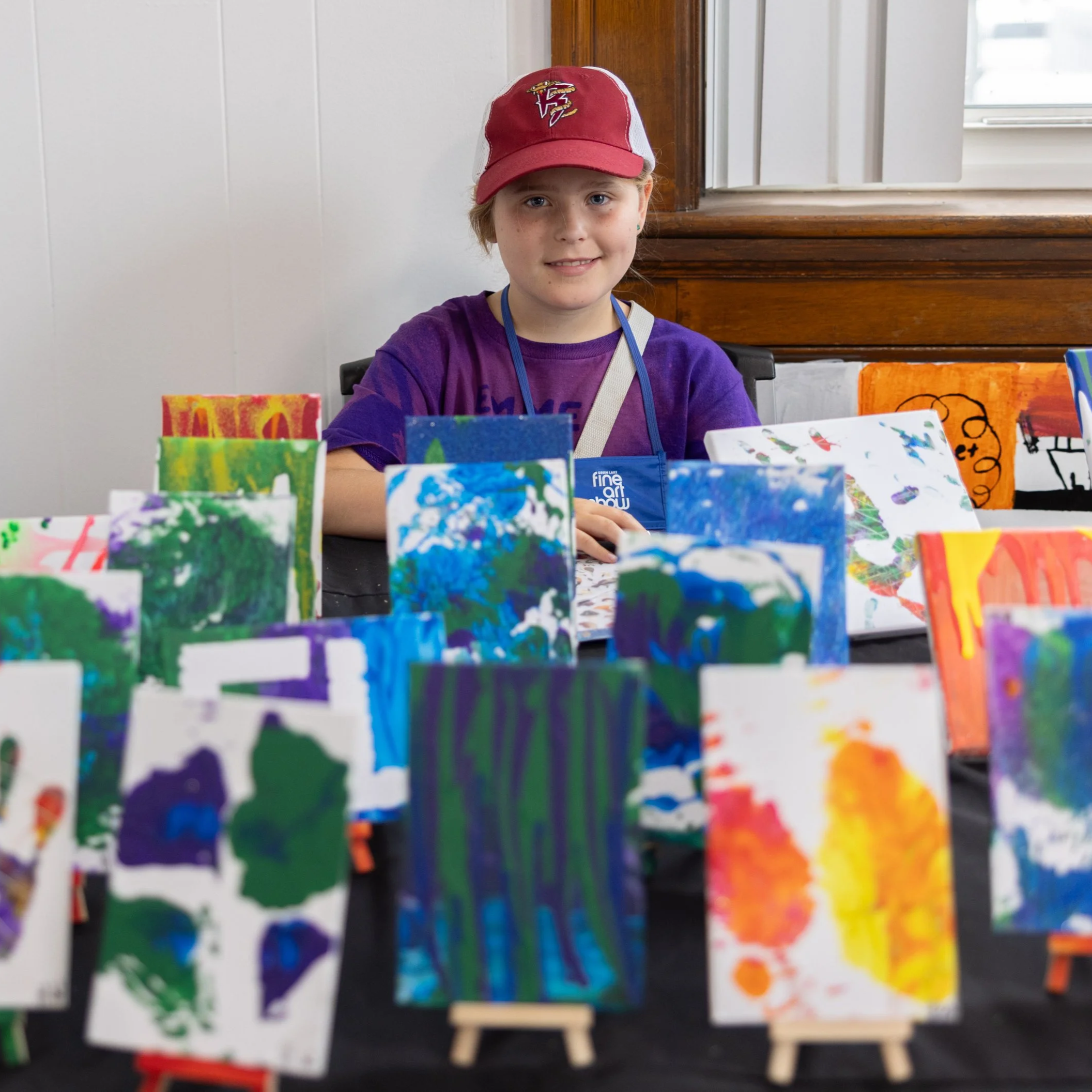 A young artist proudly displays original paintings during the Youth Art Showcase, celebrating emerging talent, creative expression, community arts programs, and the Green Lake Fine Art Show.