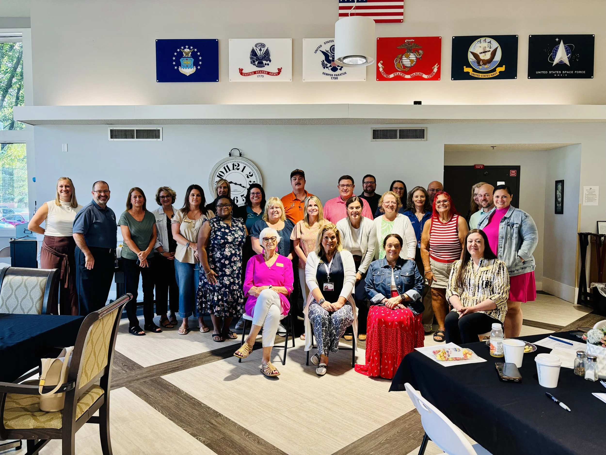 A group of diverse people, mostly women, standing and sitting together inside a building with decorated walls featuring military flags and insignias. The group is smiling and posing for a photo, with tables and chairs in the foreground.