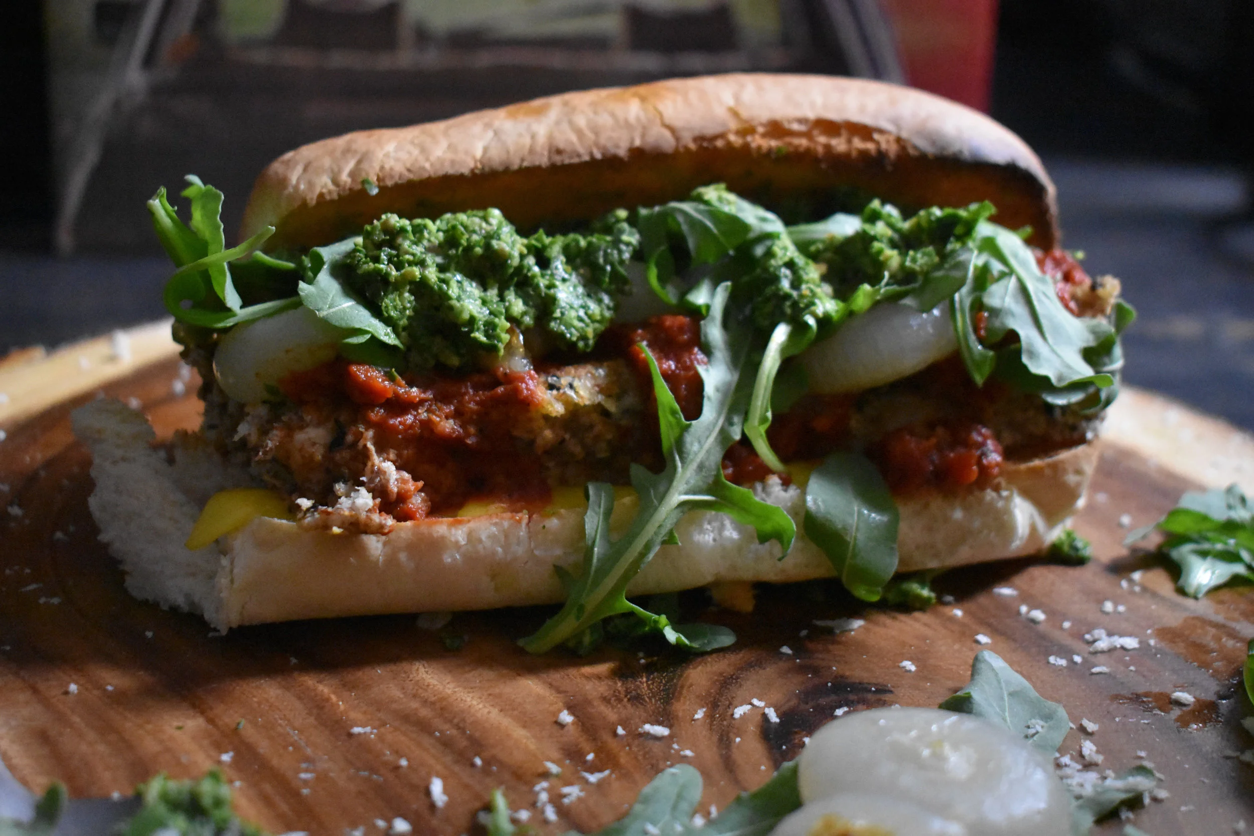 8. To finish the sandwich, add bread sliced open and a few 1/2 slices of vegan cheese to oven and broil at 525F for 5-6 minutes. Then layer on cutlets, tomato sauce, onions, pesto, and arugula. 