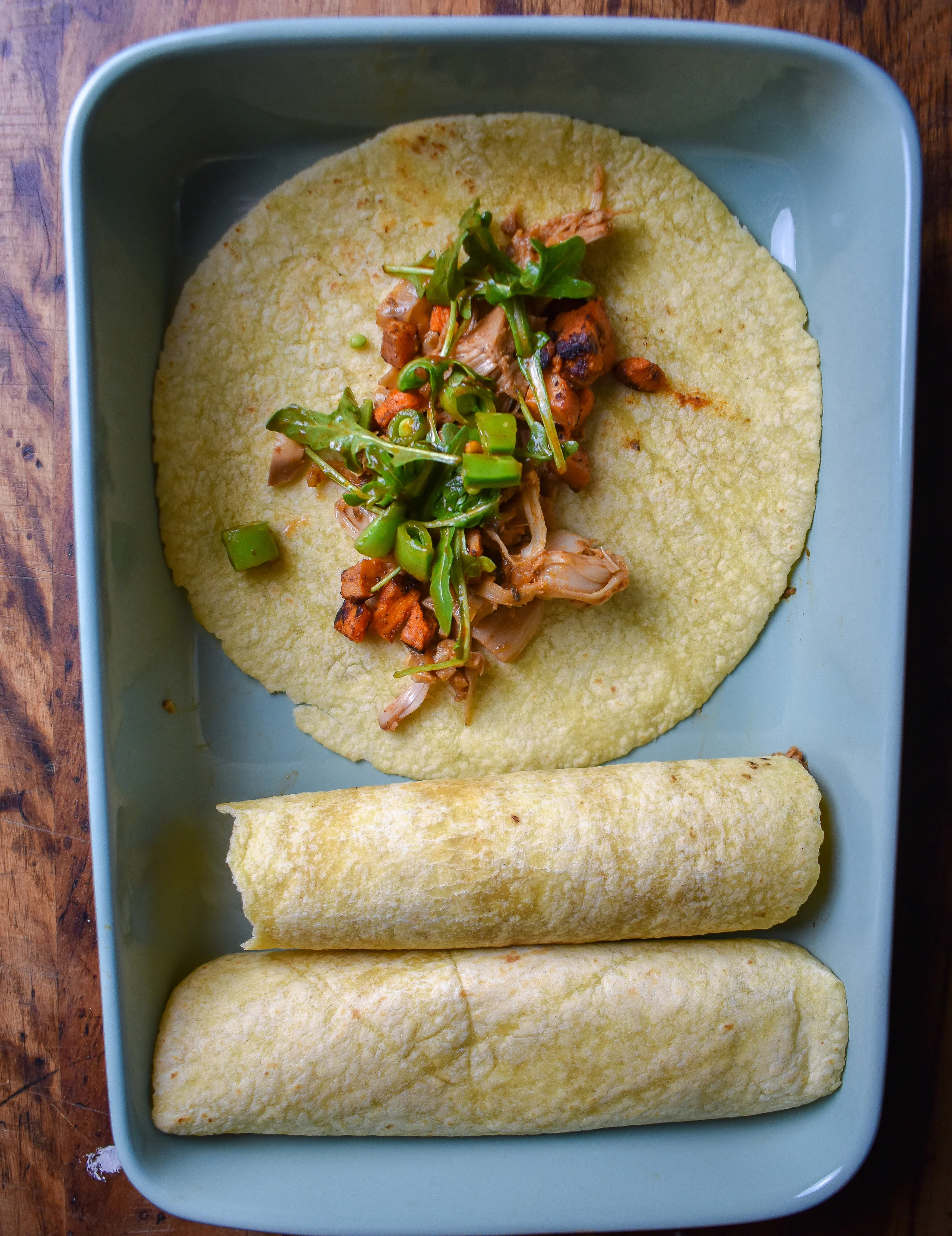 7. To form enchiladas, add carnitas to a corn tortilla, followed by potatoes, and then a handful of arugula mixture. Roll tight and place in casserole pan.