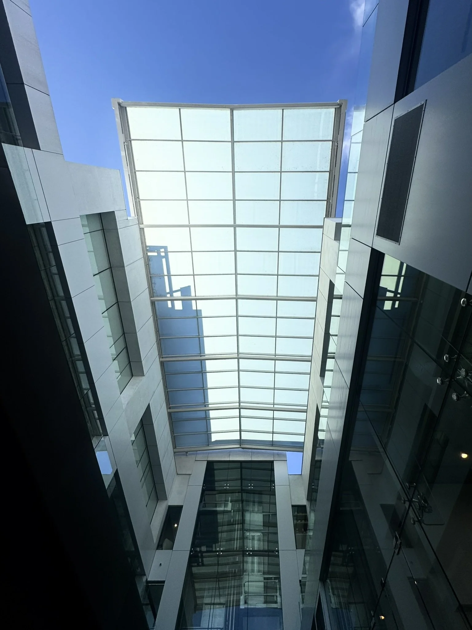 Photo of a modern building with a large glass ceiling allowing natural light to illuminate the interior, taken from the ground looking upward.