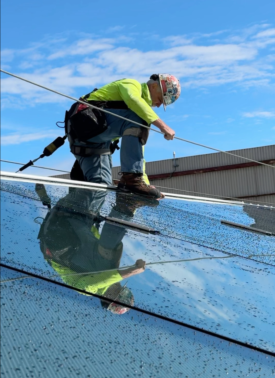 A worker balancing on a ridge skylight, wearing a helmet and bright yellow safety jacket, installing a skylight on a sunny day.