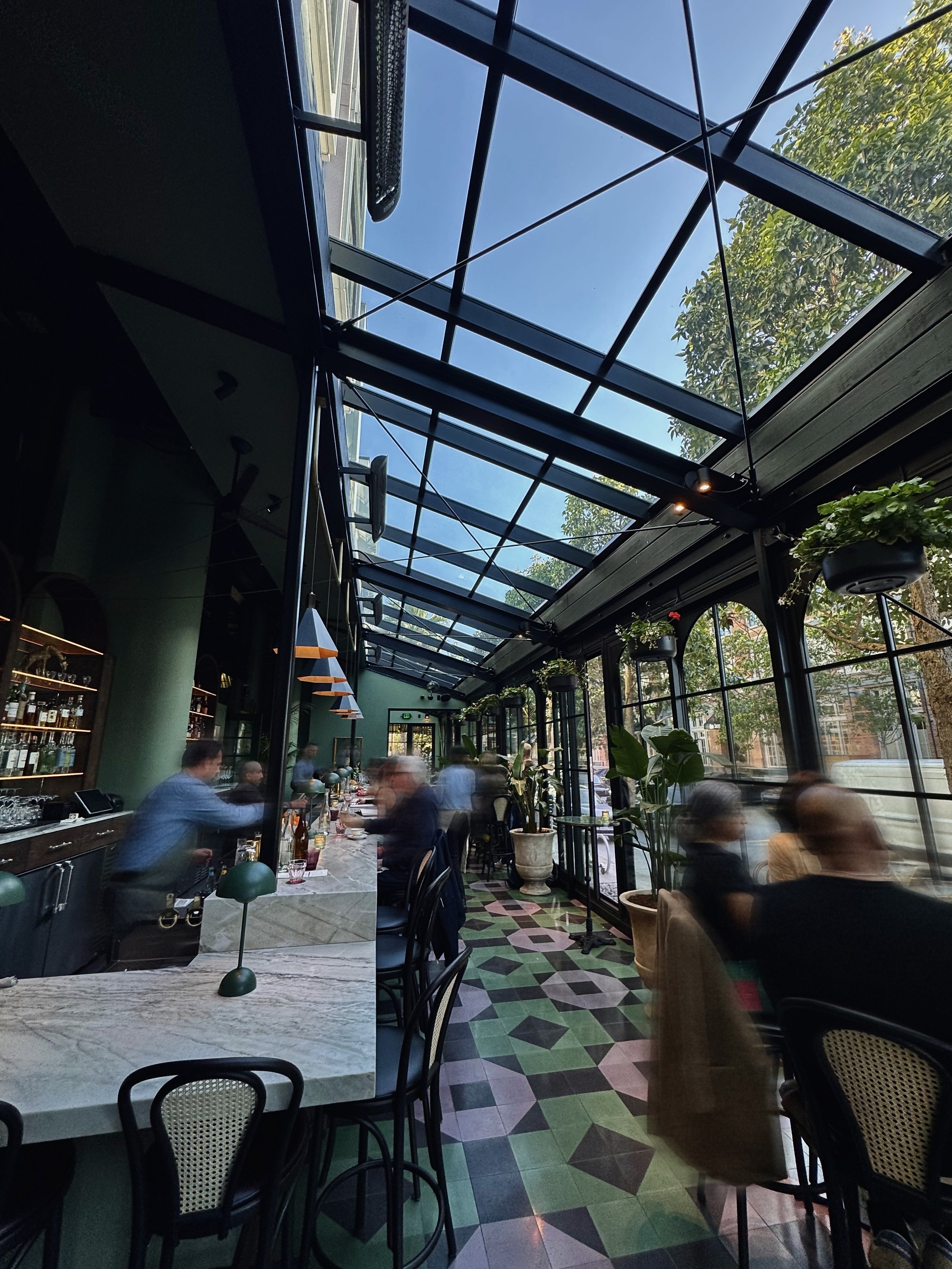 Interior of a restaurant with large skylight ceiling, green walls, dining tables, chairs, and blurred patrons dining. Outside trees visible through windows.