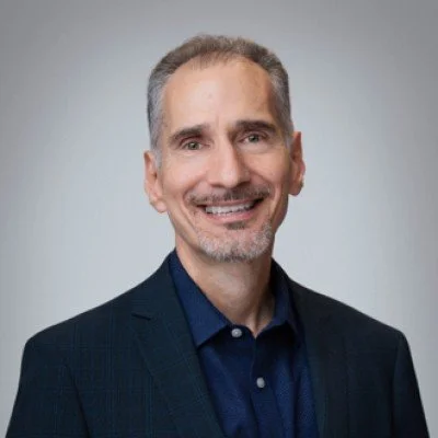 A smiling middle-aged man with short gray hair and a beard, wearing a dark suit and blue shirt, in front of a plain gray background.