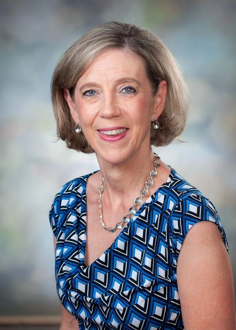 A portrait of a smiling middle-aged woman with short blonde hair, wearing a blue geometric patterned top, silver jewelry including earrings, necklace, and bracelet, with a blurred neutral background.