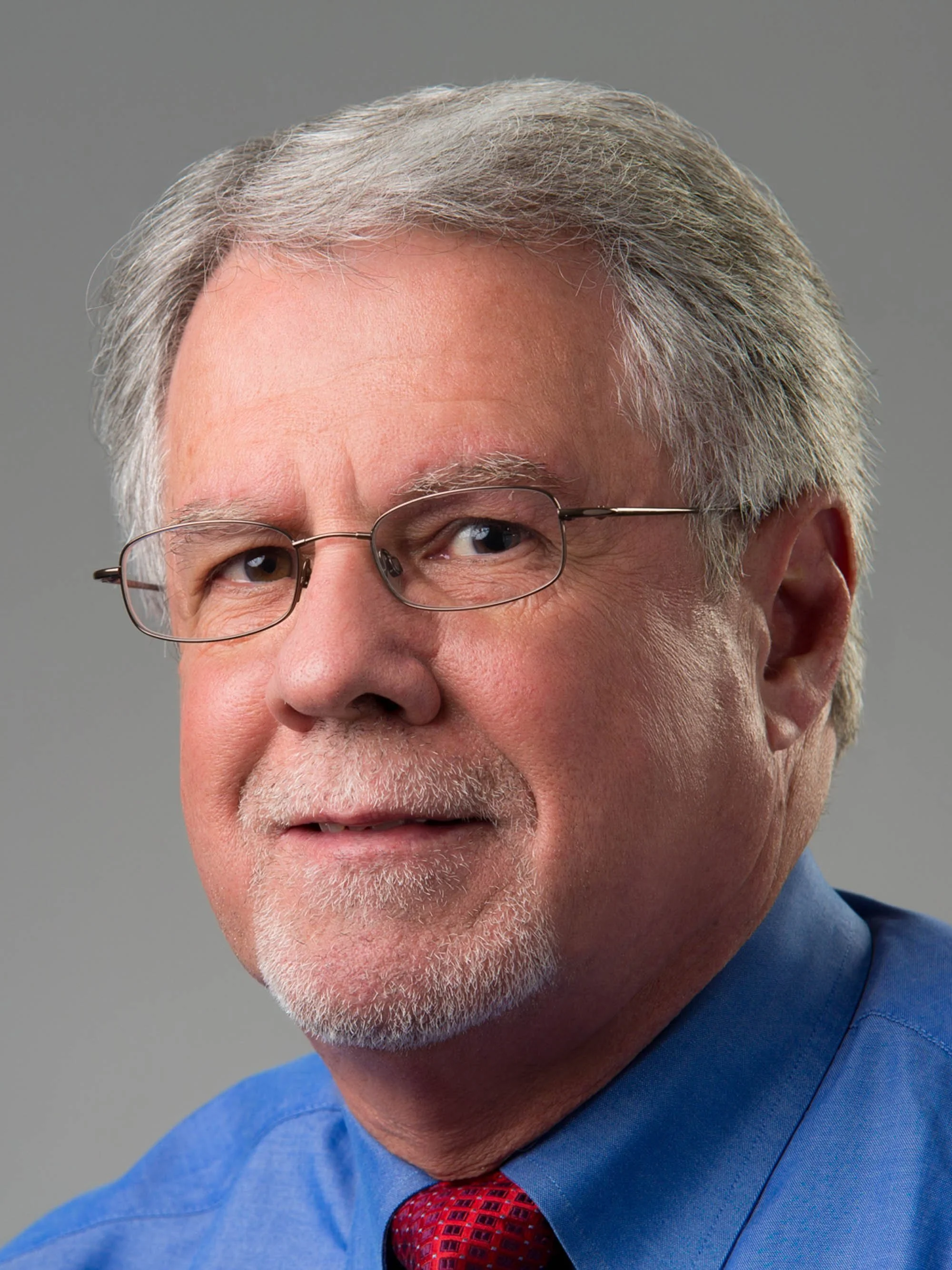 Close-up portrait of an older man with gray hair, glasses, and a short beard. He's wearing a blue shirt and a red tie, looking at the camera with a slight smile, against a gray background.